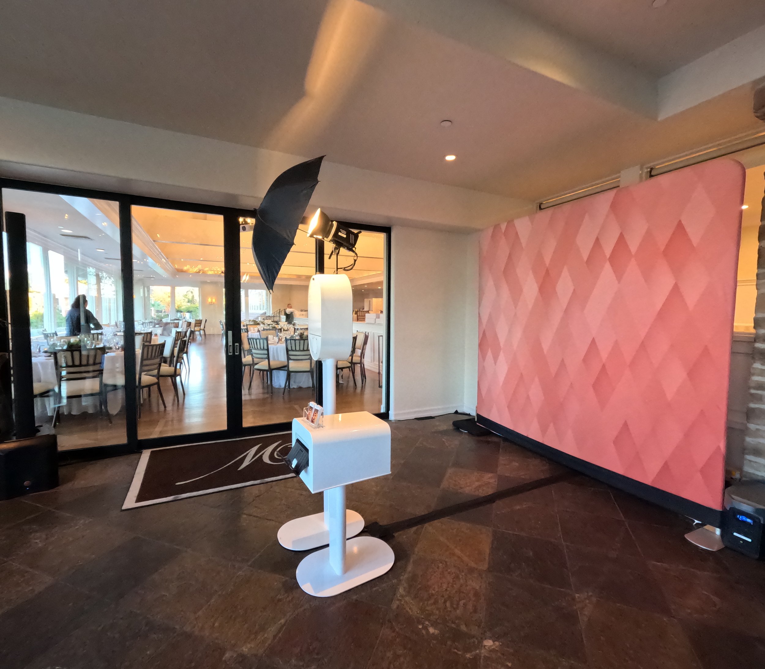 Photo of a photo booth setup in an indoor venue with a pink geometric backdrop, studio lighting equipment, and a white photo stand in front of a doorway leading to a restaurant or event space.