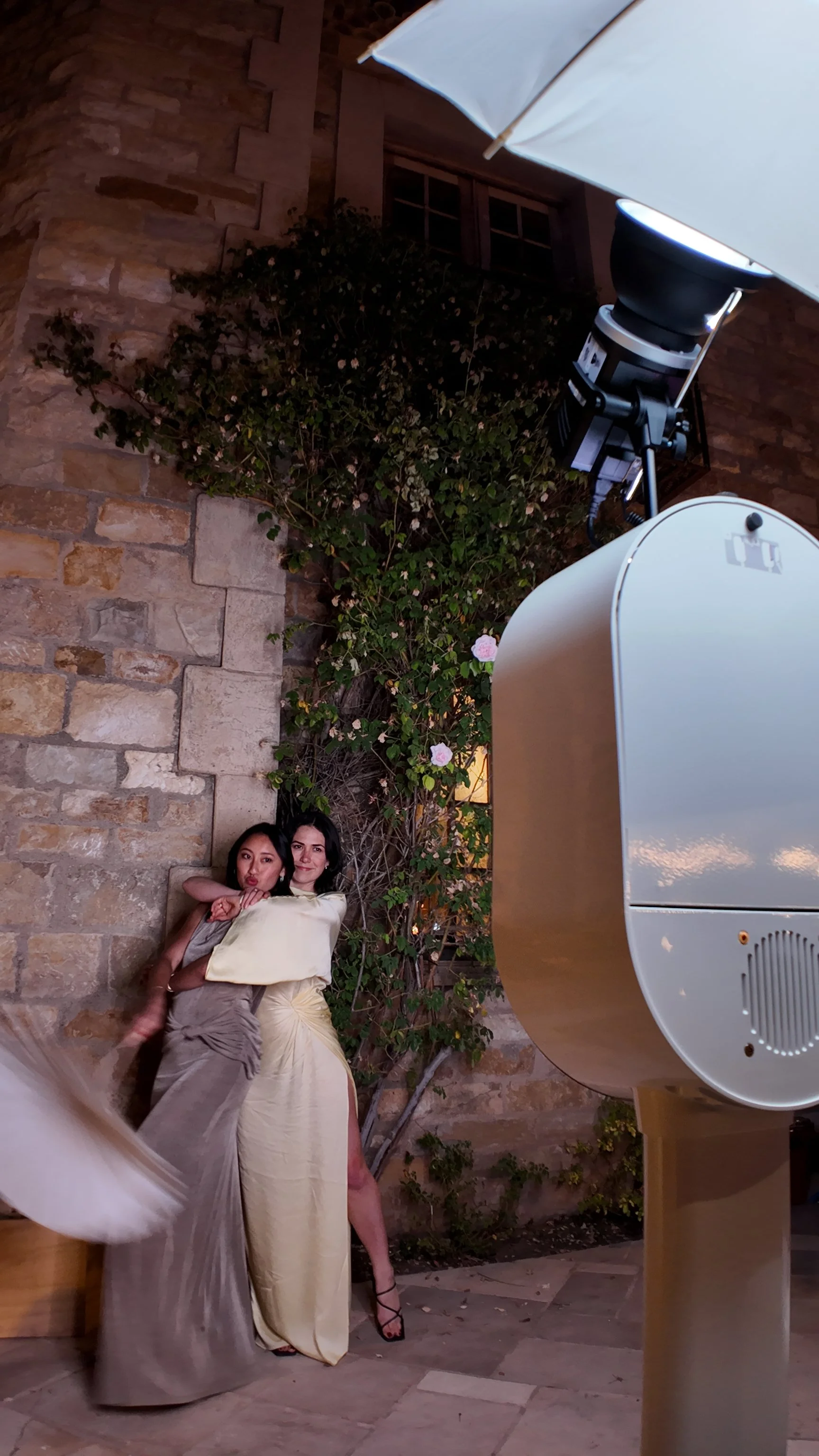 Two women in elegant dresses posing in front of a stone wall with flowering plants, taking a photograph with a large camera and lighting equipment outdoors at night.