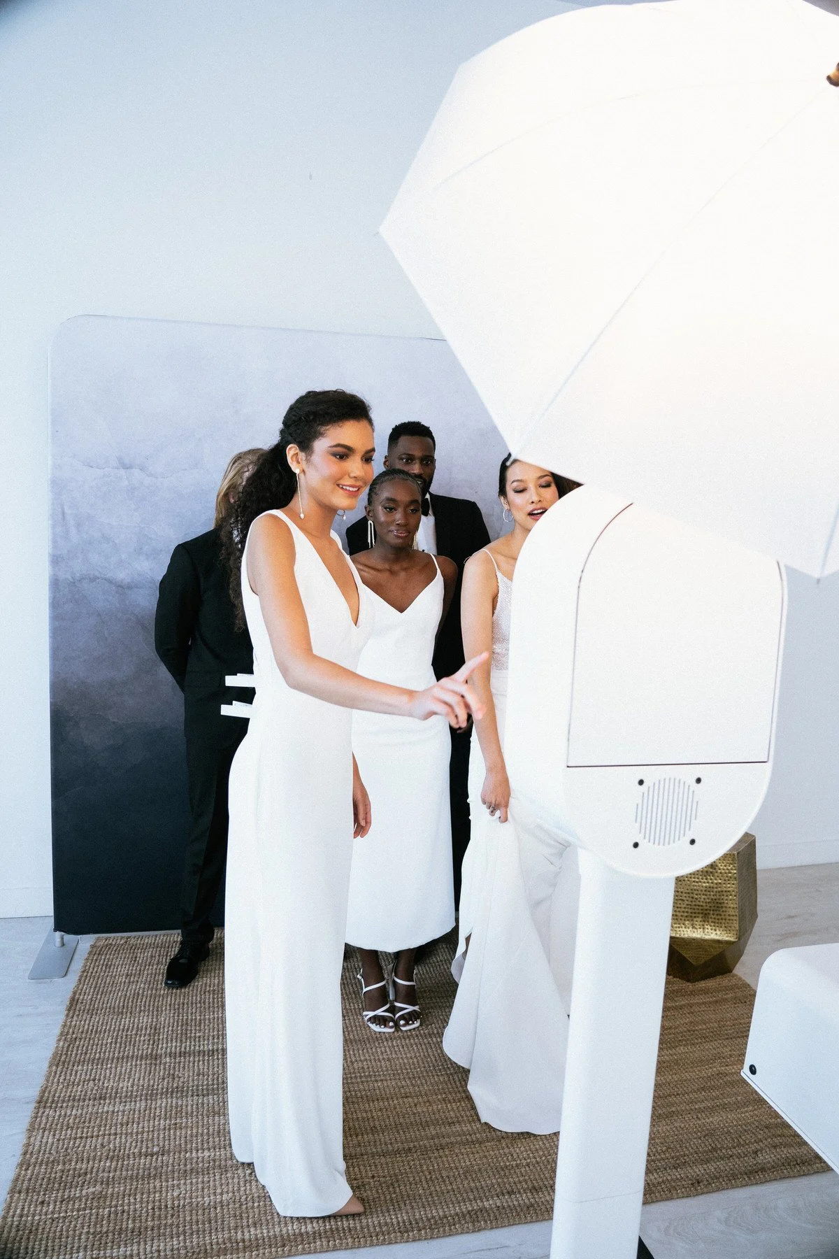 A group of five diverse women and one man in formal attire looking at a photo booth camera and smiling.