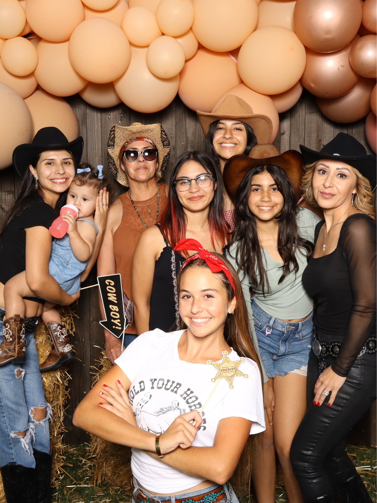 A group of women and a young girl dressed in cowboy and western-themed clothing, posing for a photo with balloons and a wooden backdrop.