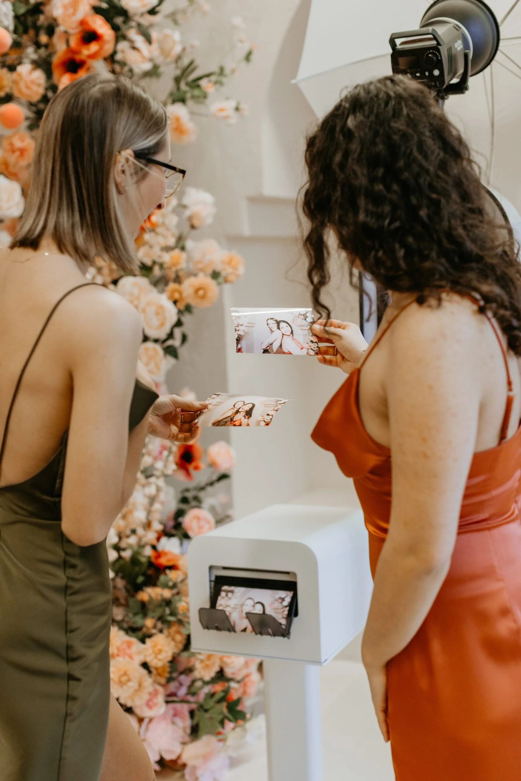 Two women in elegant dresses, one in olive and the other in rust, stand in front of a floral backdrop at a photo booth, holding printed photos of themselves during an event.