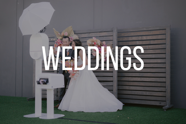 A bride and groom at a wedding photo booth with a gray wall and wooden fence background, floral arrangements, and photography equipment.