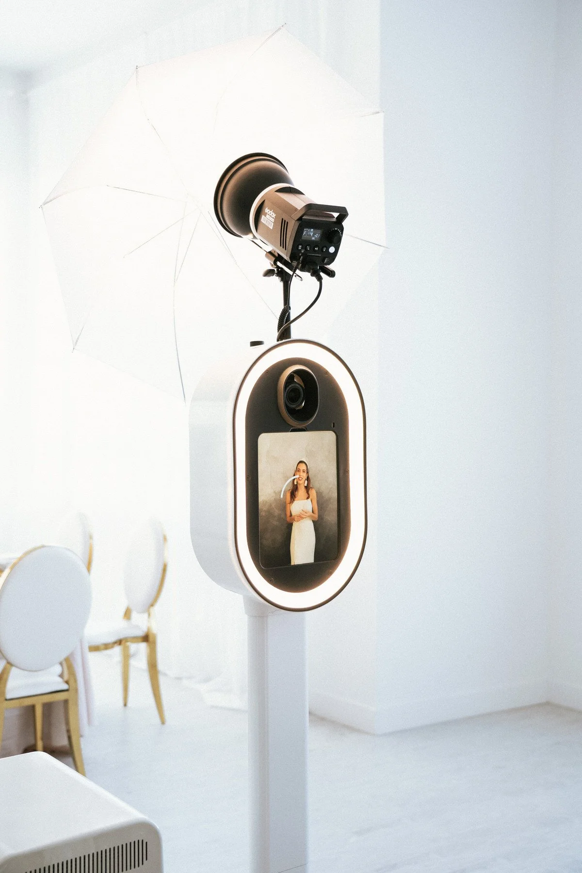 A camera and lighting setup for taking photos of a woman, who is displayed on the camera's screen, in a white studio with chairs and a white umbrella reflector.