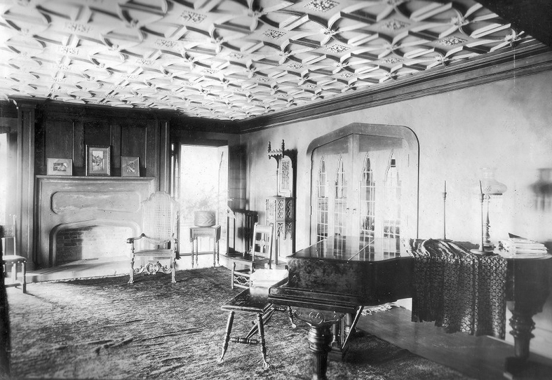  Black and white photo of lavishly decorated room, with piano in foreground and empty fireplace at the far side 