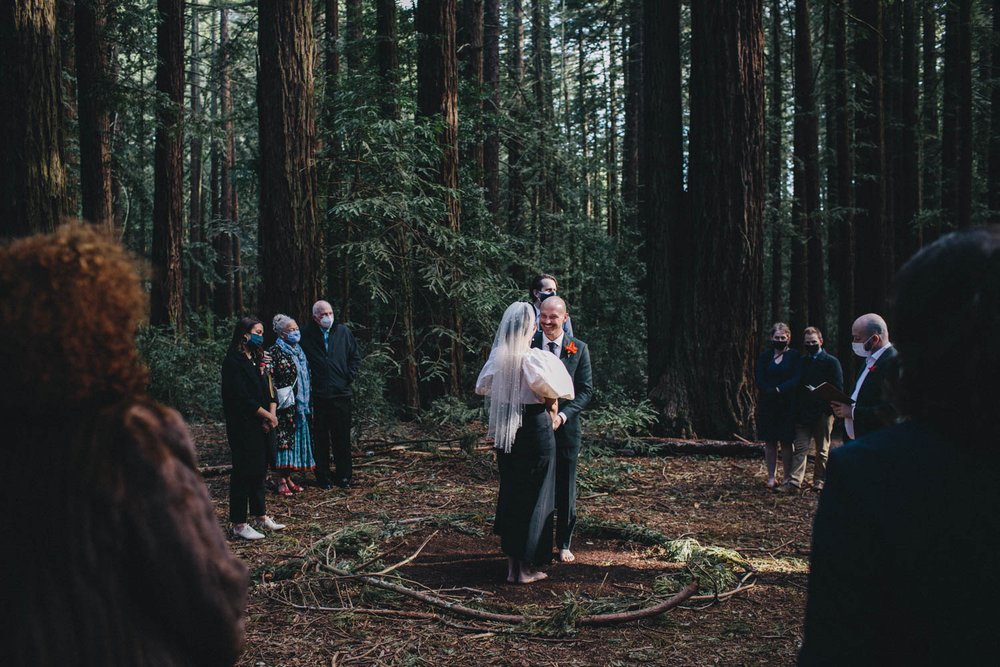 a couple stands in the center of a circle of friends and family during their wedding ceremony in a redwood grove at joaquin miller park