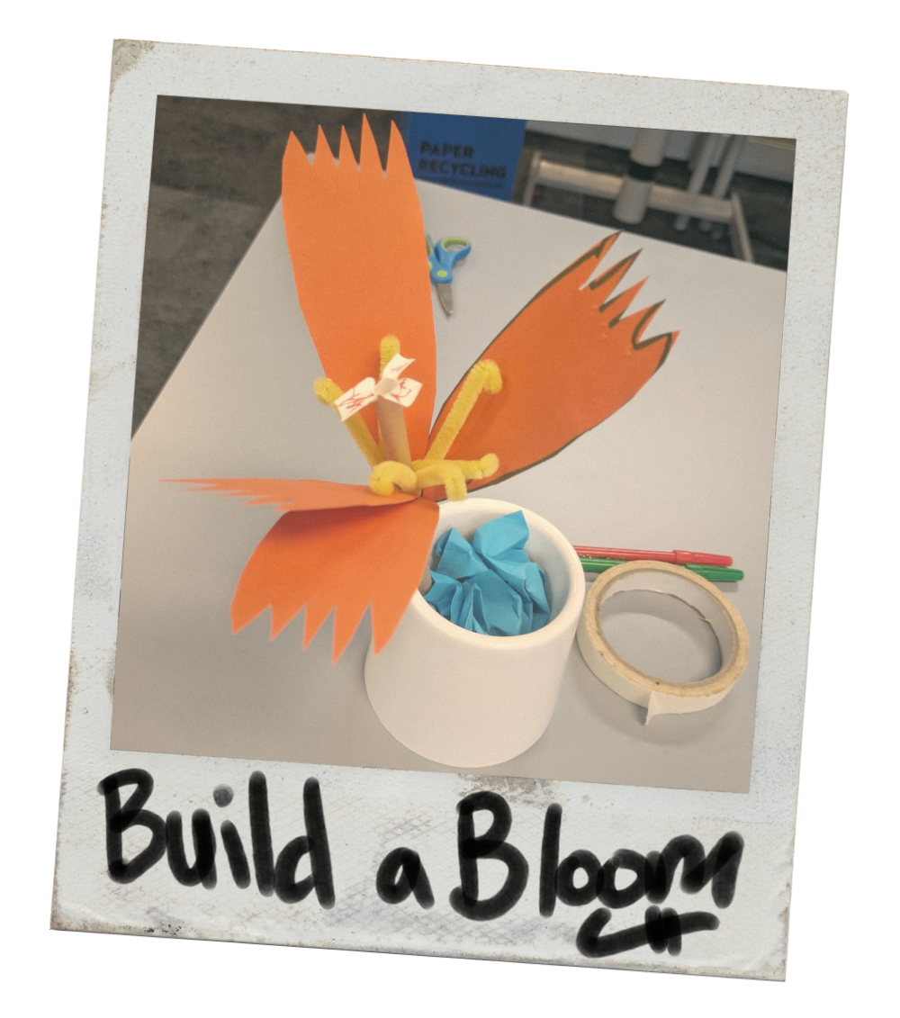 A  paper flower with orange petals, yellow pipe cleaners for the stamen, and a straw for the pistil. It is placed in a white cup filled with blue tissue paper. The project is labeled "Build a Bloom" in black marker on a white background.