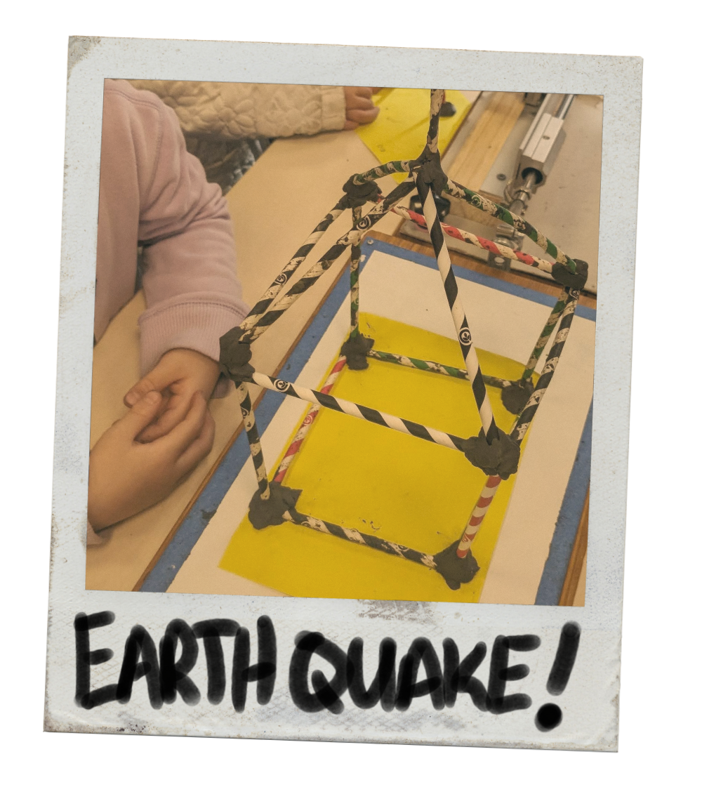 A tabletop earthquake simulator, testing a building made by a student from paper straws and plasticine. The word "EARTHQUAKE!" is written at the bottom of the photo.