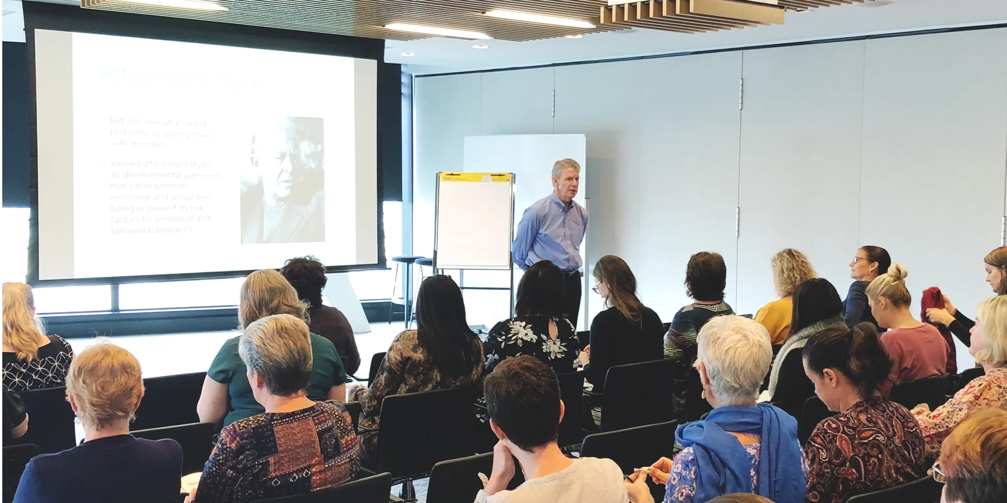 Image: Man standing in front of an audience to the right of a projector screen. Audience have their back to the camera.