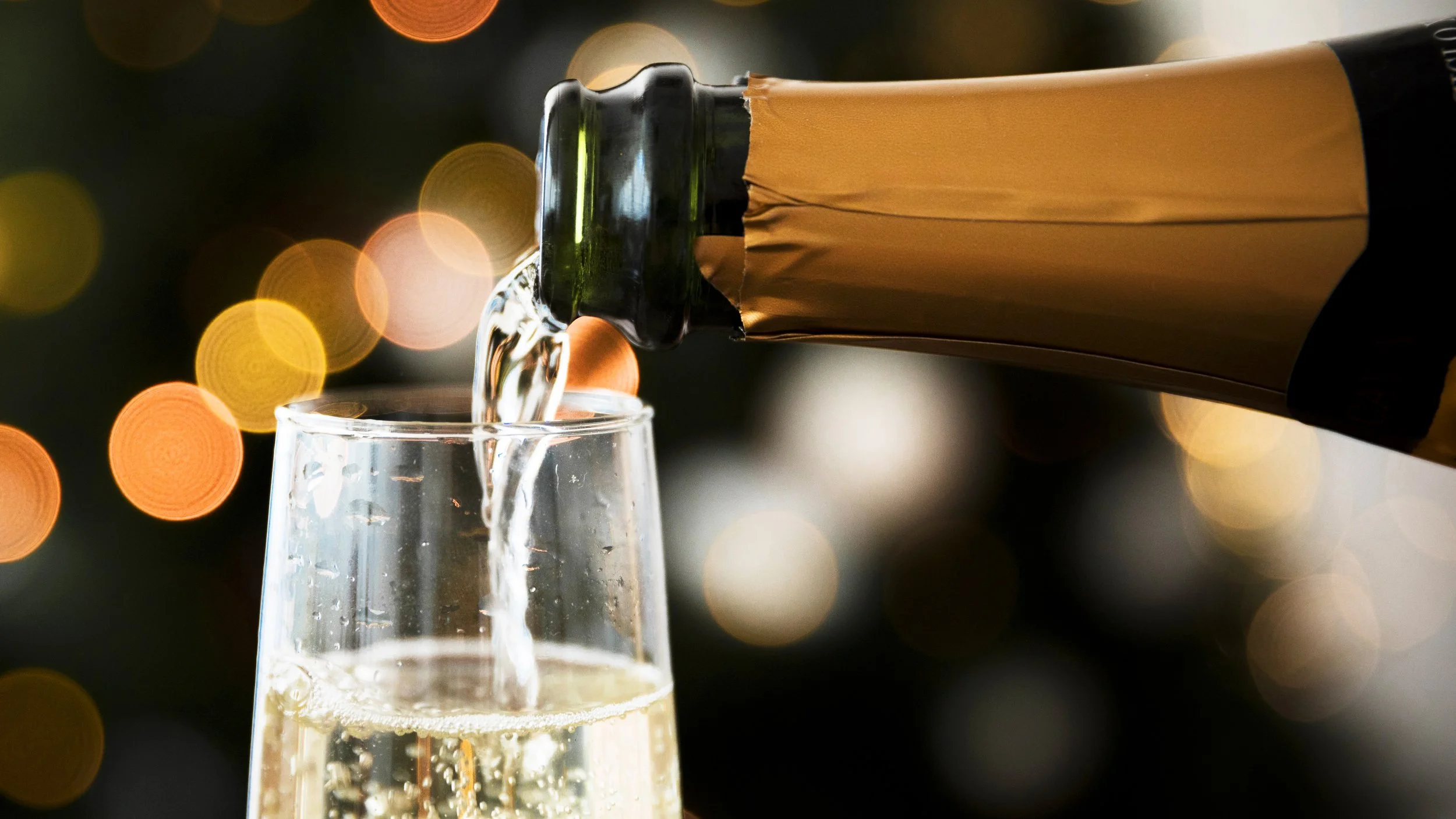 A bottle of champagne is being poured into a champagne flute, with a backdrop of blurred festive lights.