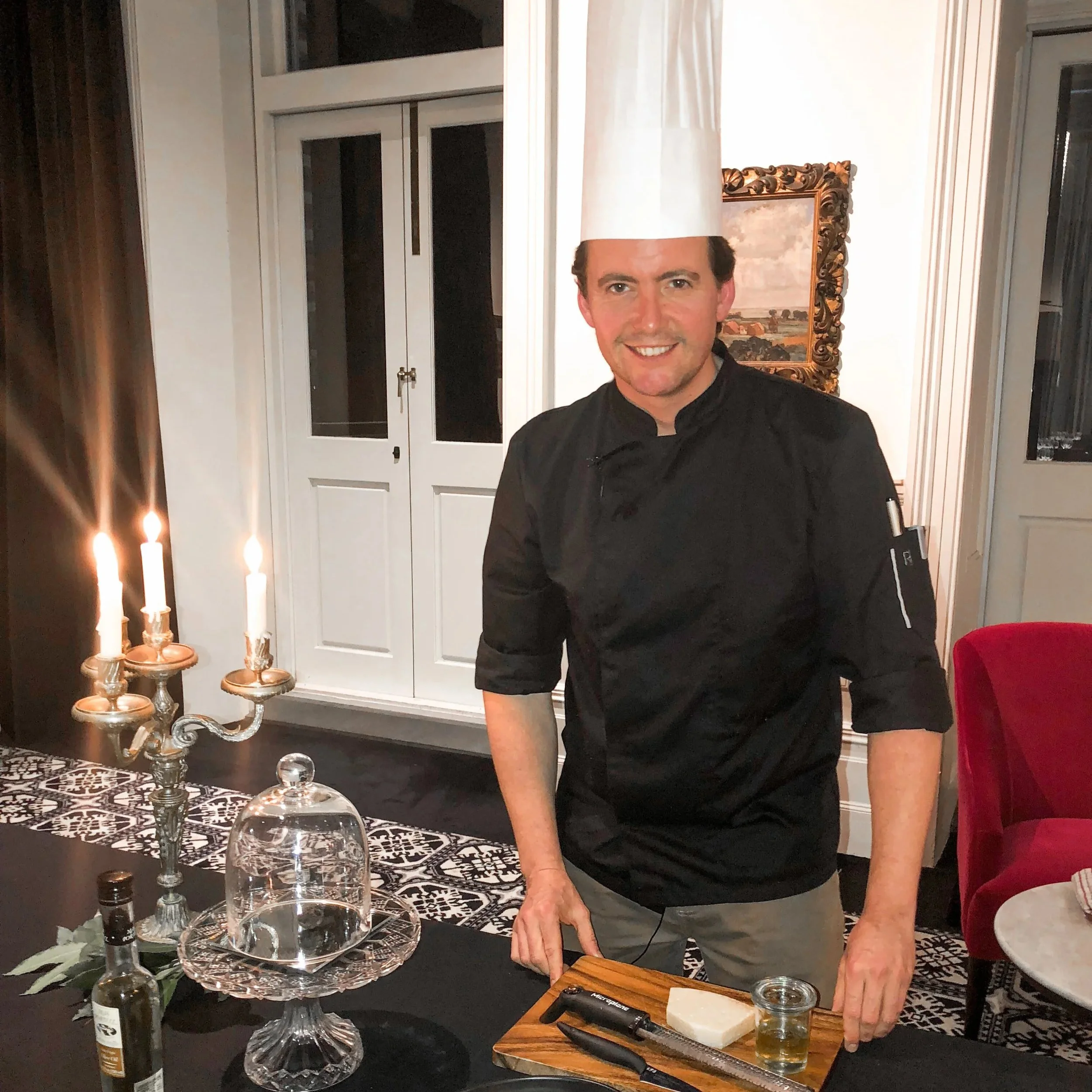 A male chef stands in a restaurant kitchen, wearing a white chef's hat and black uniform, smiling at the camera, with a cheese and wine setup on a table in front of him.