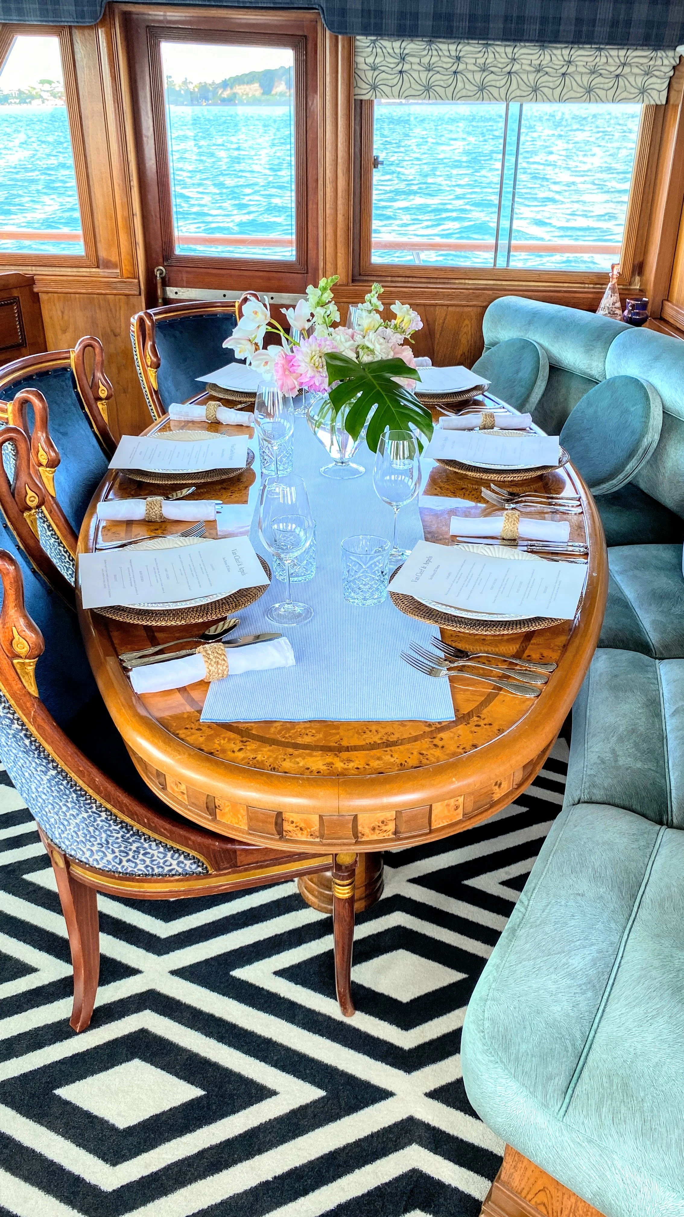 A dining table set for a formal meal with six placemats, white napkins, and menus, decorated with a central vase of pink and white flowers, in a room with large windows showing a body of water outside.