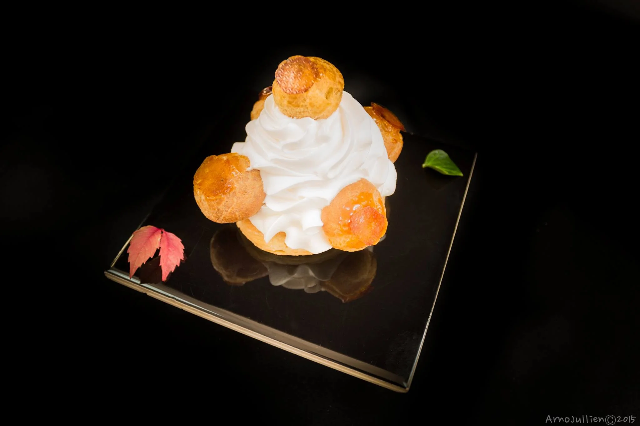 A Saint Honoré cake with cream puffs and whipped cream on a reflective surface, accompanied by a small red leaf and a green leaf.