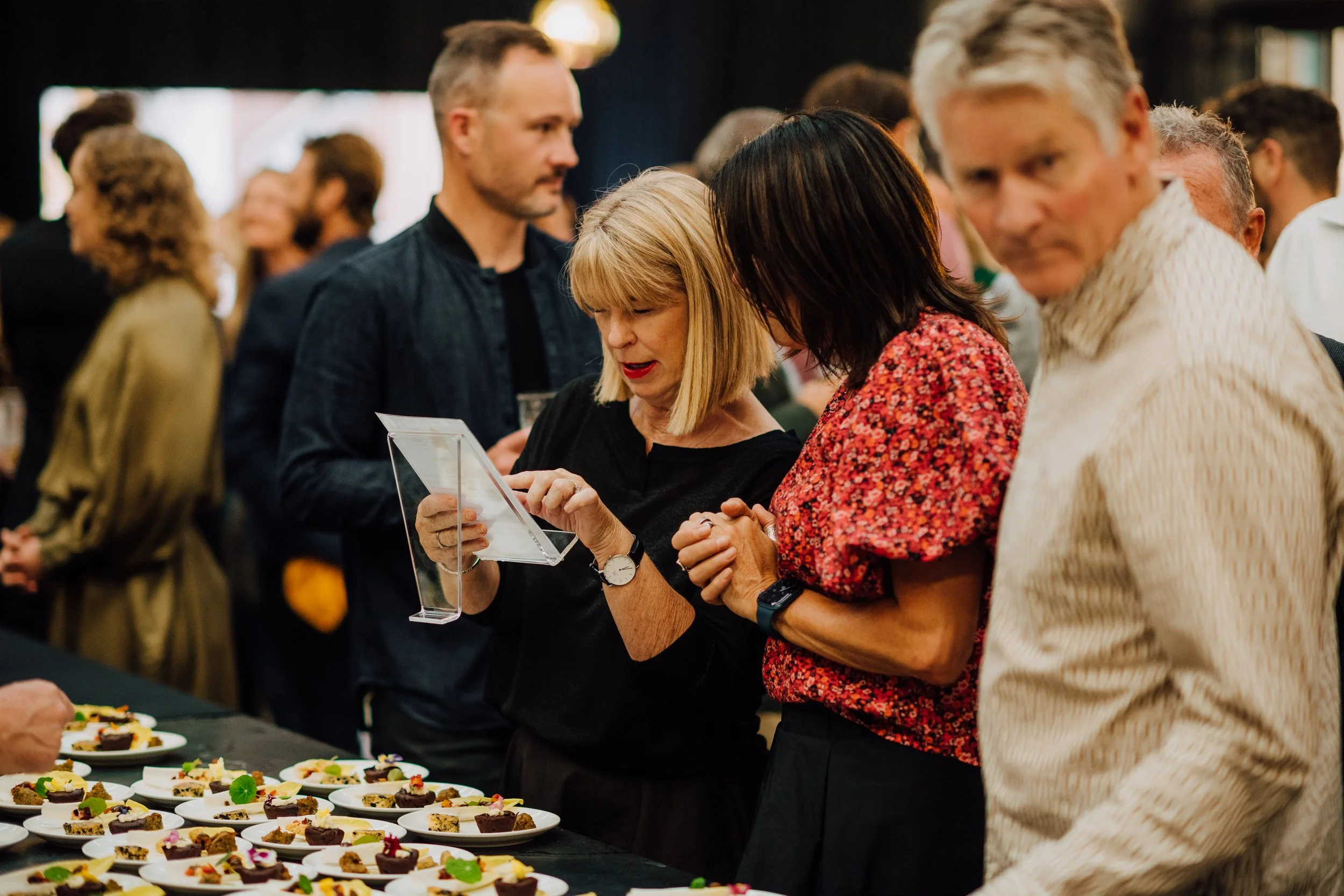People at an event examining food on plates, with a woman holding a menu card.