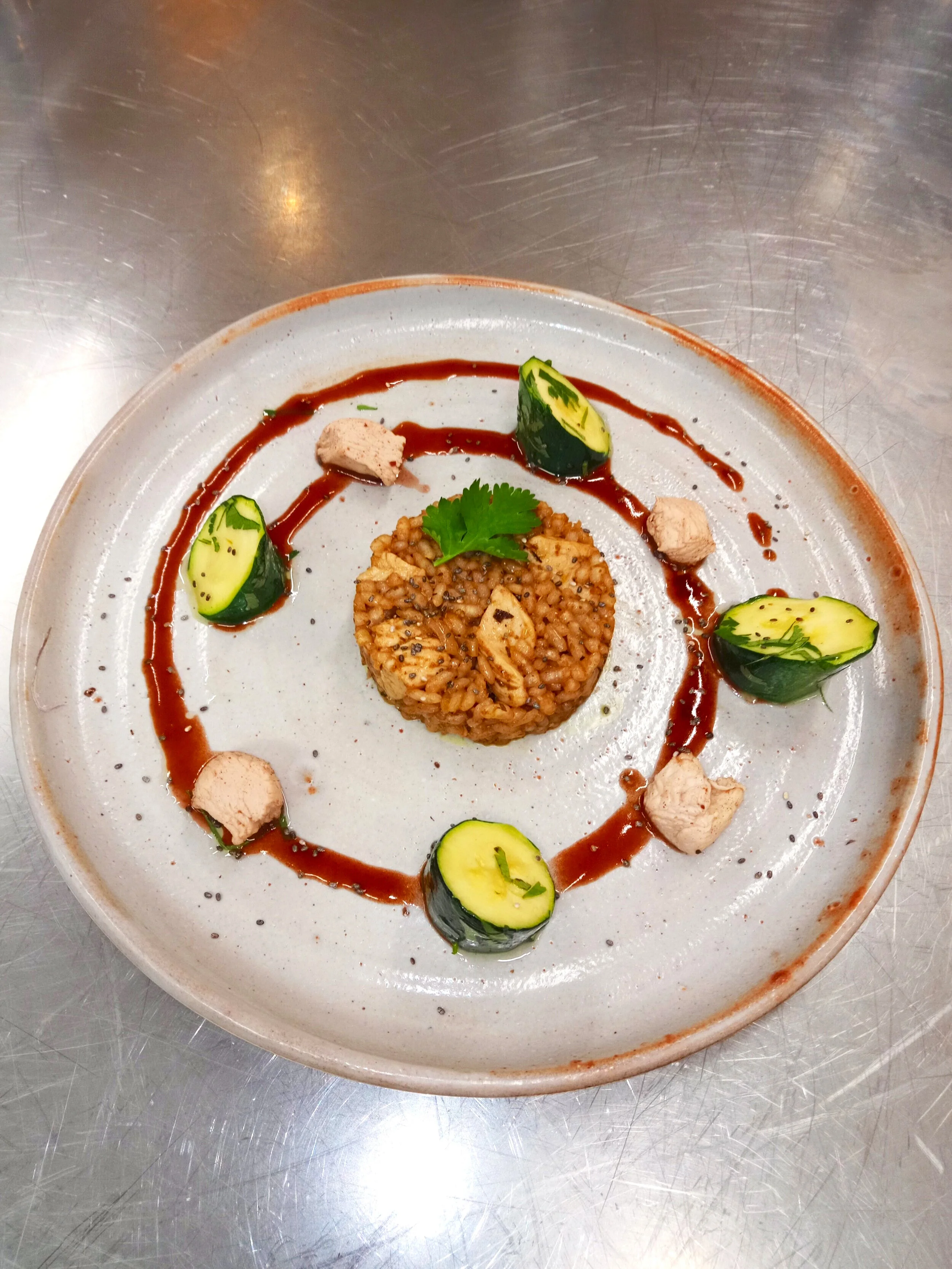 Gourmet dish featuring a central mound of seasoned rice, surrounded by decorative sauces, zucchini slices, and small pieces of meat, garnished with a fresh herb on a ceramic plate.