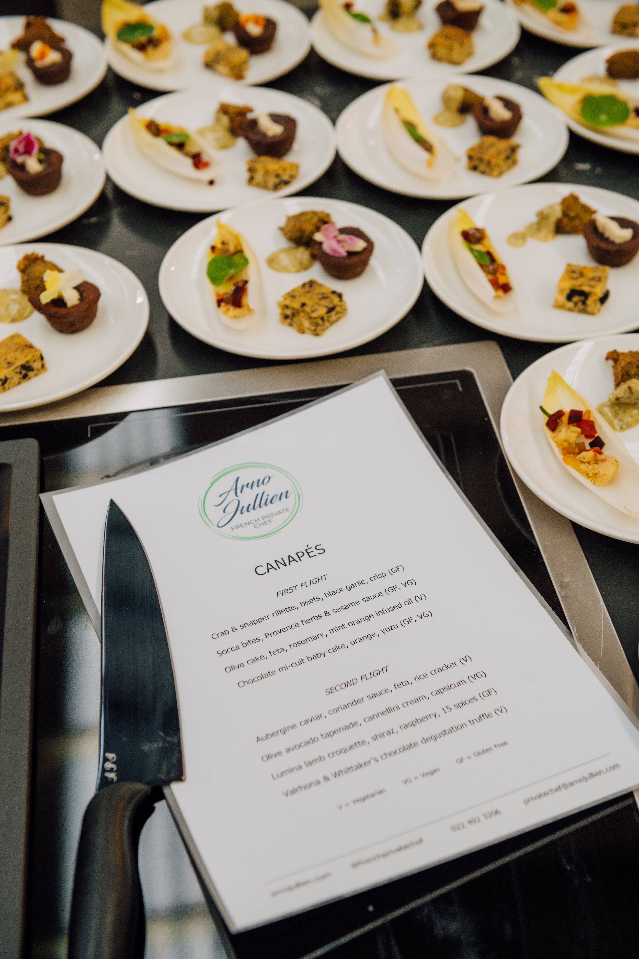A table with multiple small plates of canapés, each containing a variety of appetizers like stuffed endives and cubes. A menu titled 'Canapés' lists items such as crab & snapper rillette, socca bites, and olive cake. A knife is placed beside the menu