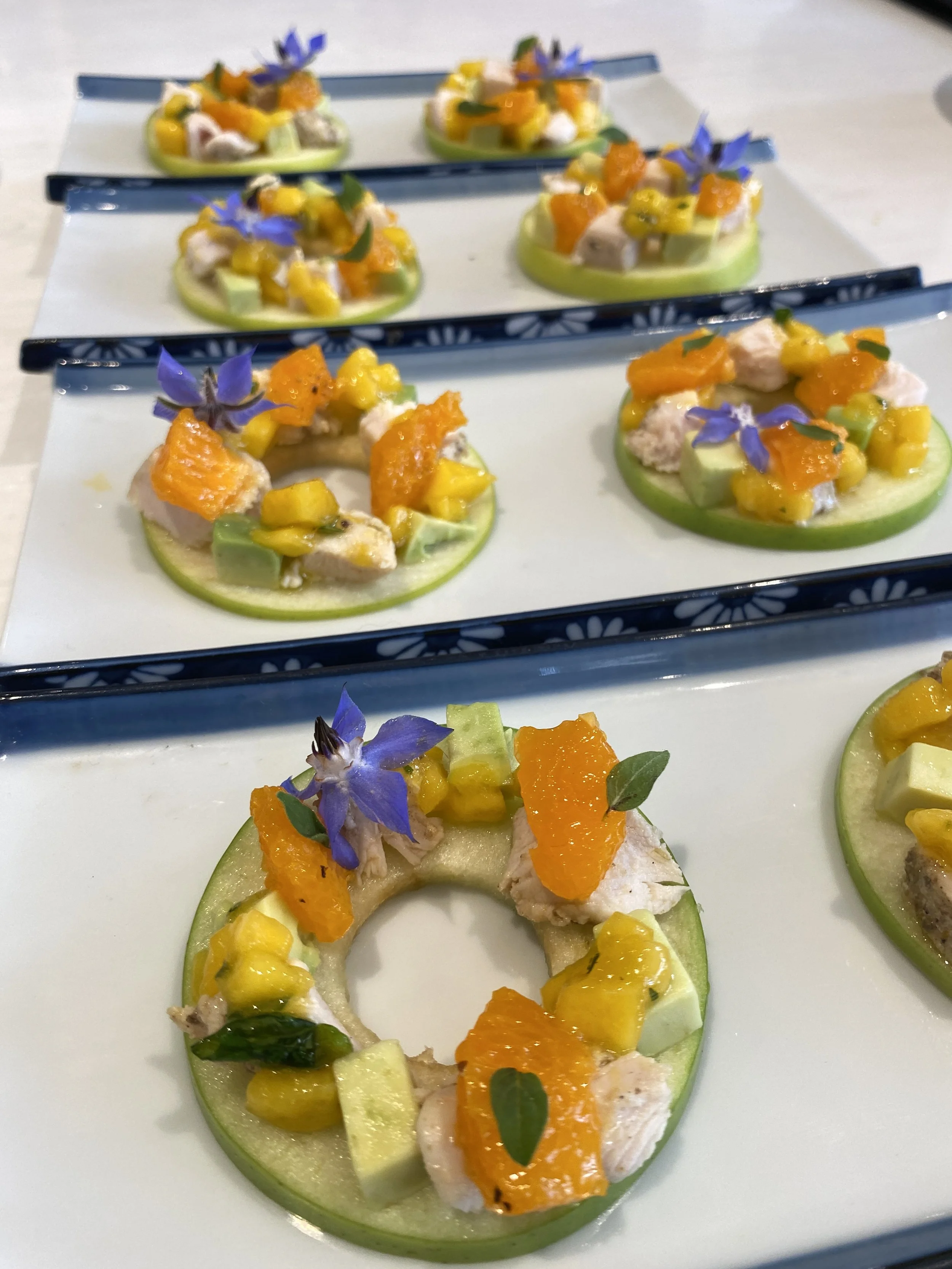 Appetizer with apple rings topped with chicken, orange segments, yellow fruit, green garnish, and purple edible flowers on a white rectangular plate.