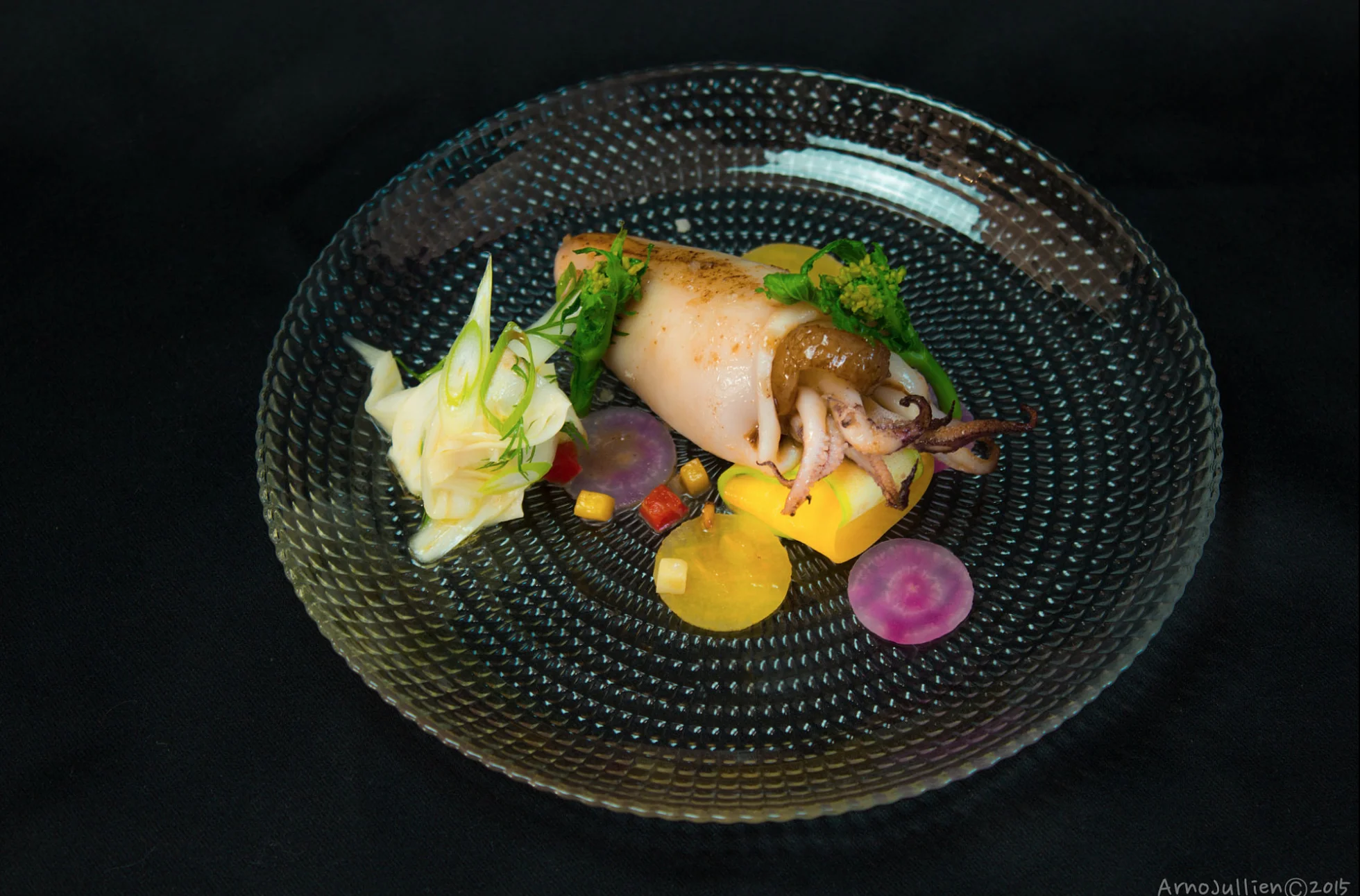 Gourmet dish featuring a stuffed squid on a glass plate, garnished with sliced vegetables, herbs, and sauce, arranged artistically on a black background.