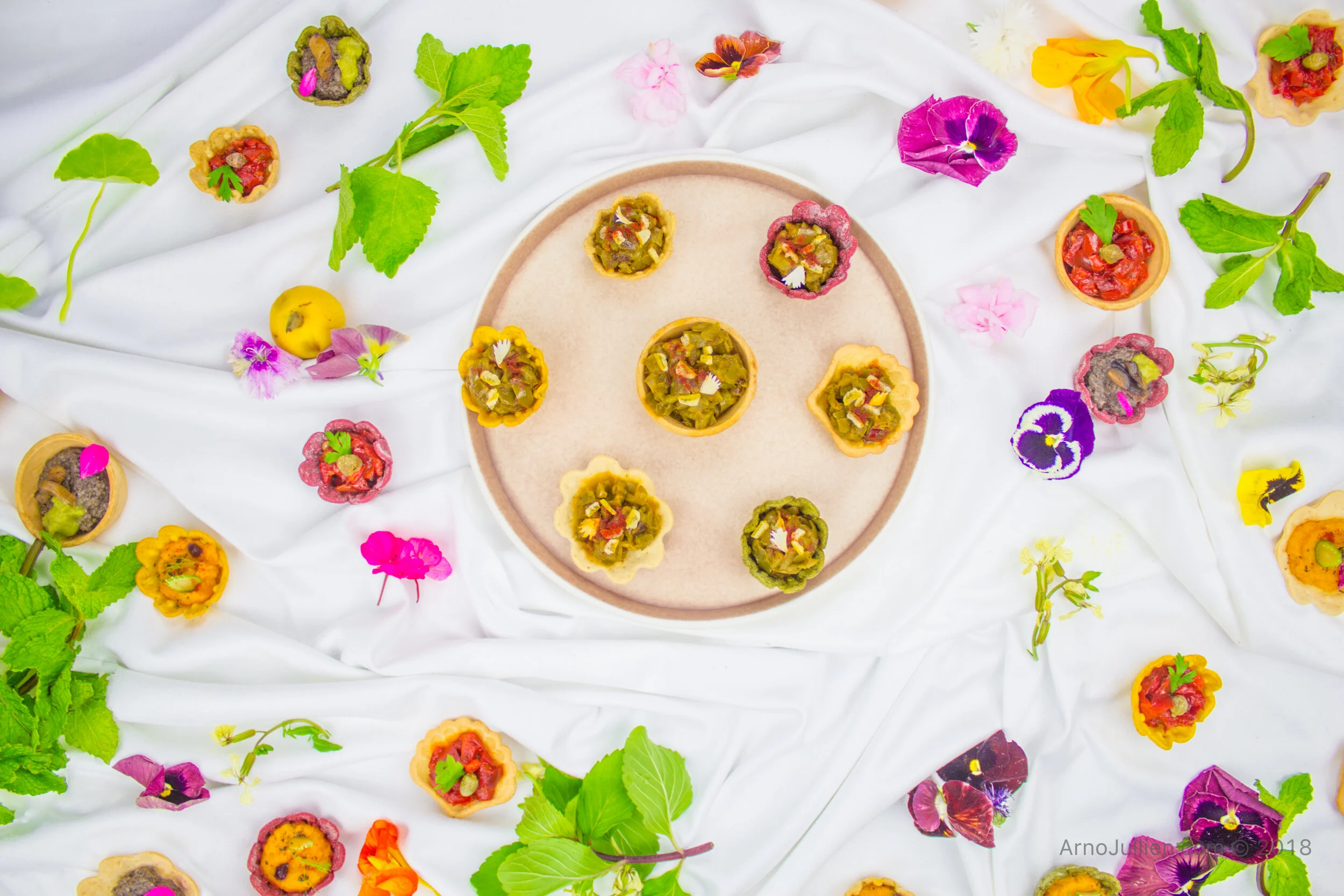 Colorful mini tarts and edible flowers arranged on a white cloth for a food-style that is elegant, colorful, seasonal and beautiful.