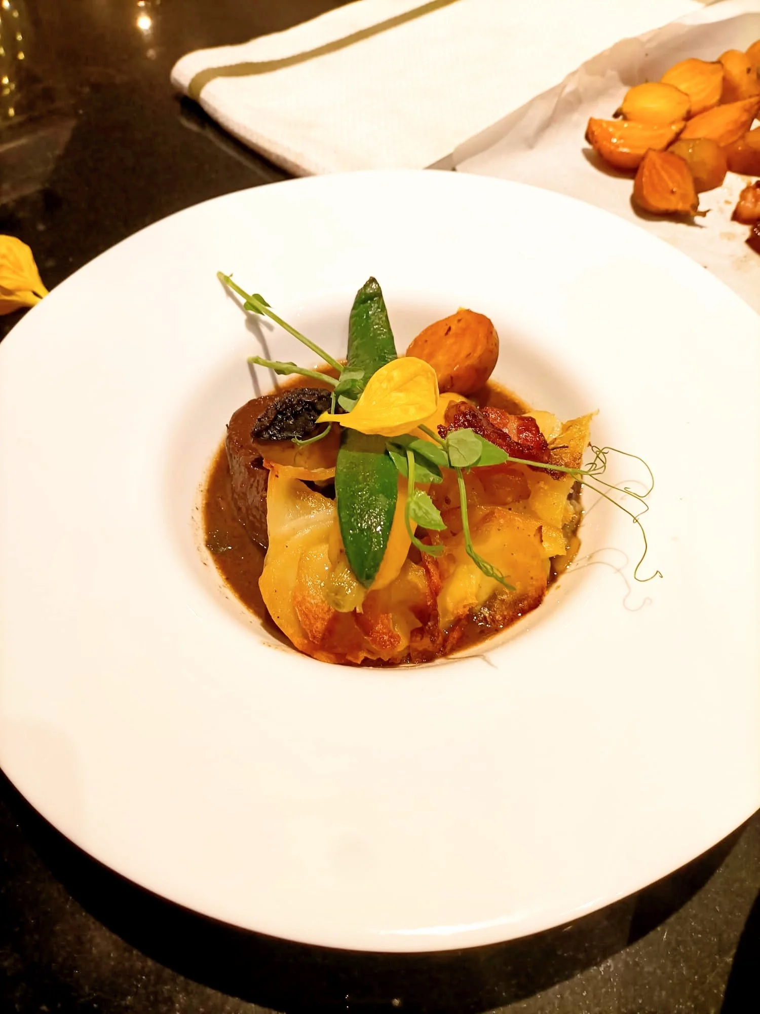 A gourmet dish featuring a piece of meat topped with various garnishes, including a yellow edible flower, green leaves, and sliced vegetables, served in a white bowl on a black table. In the background, roasted vegetables are visible on a tray.