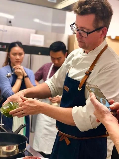 Chef conducting cooking class, pouring oil into pan, with focused students watching in kitchen setting.