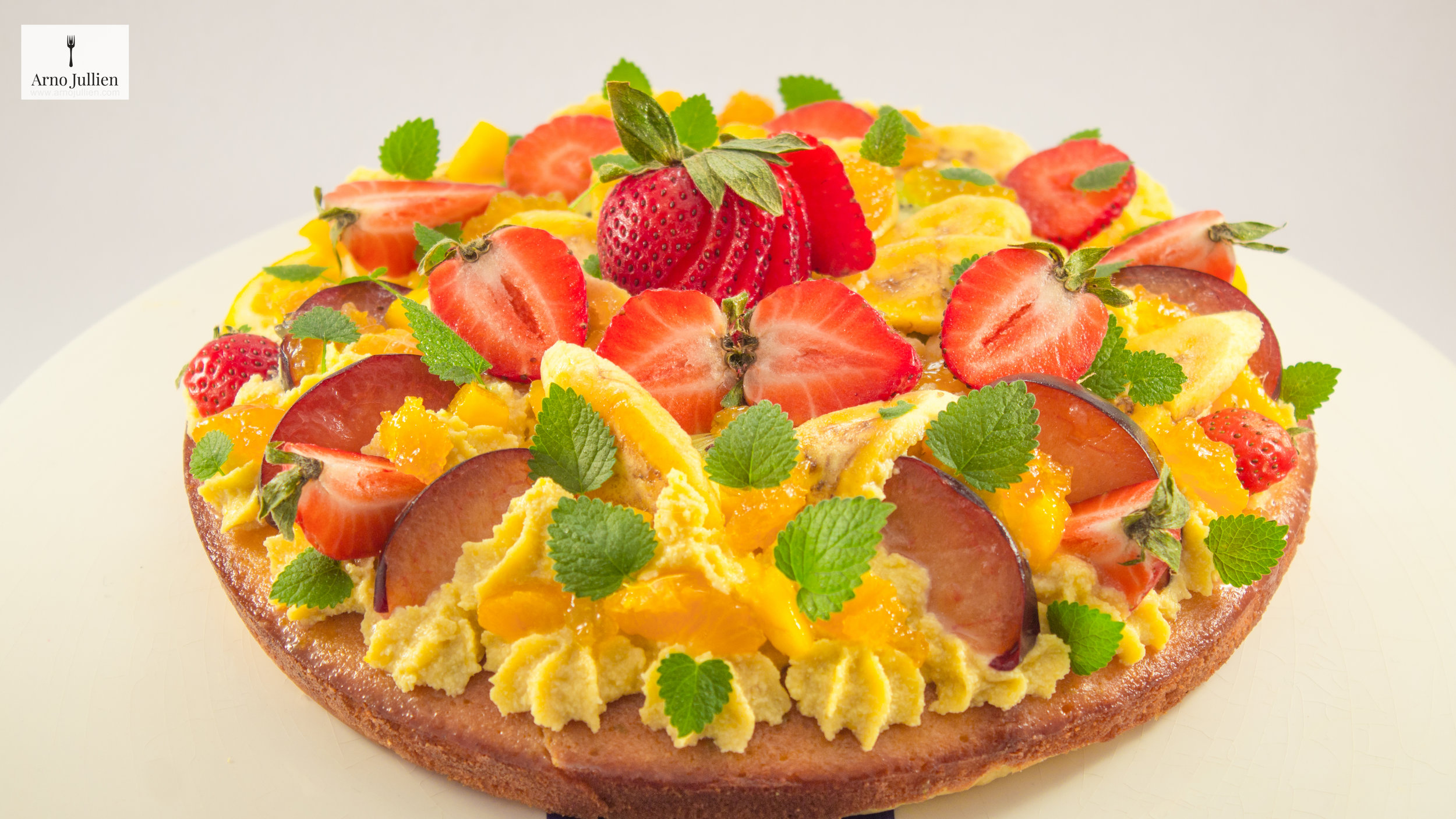 Colorful fruit tart with strawberries, plums, mangoes, and mint leaves on a light-colored cake base.