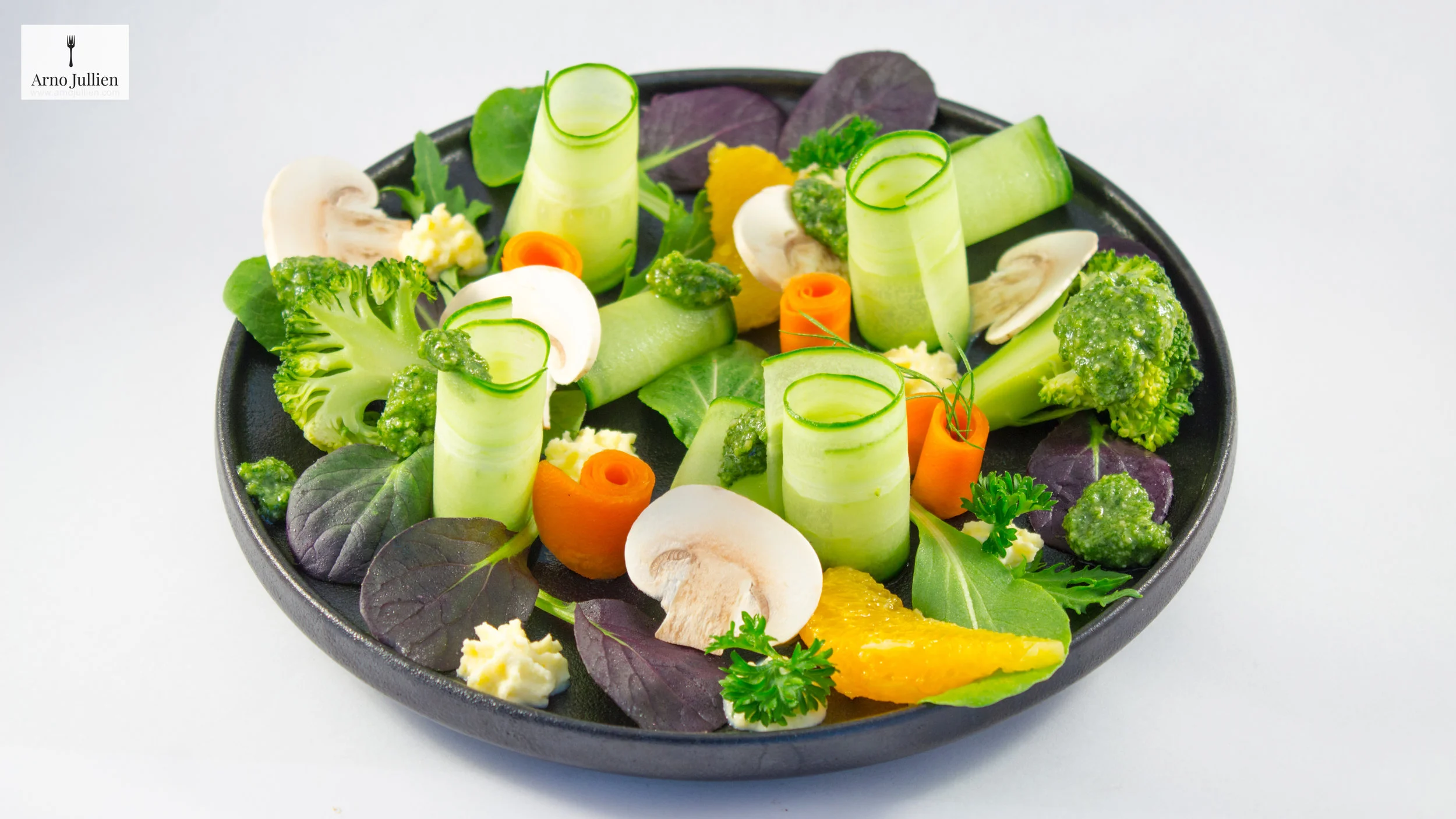 Colorful vegetable salad with cucumber rolls, carrot curls, broccoli, mushrooms, purple and green leaves, and orange slices, topped with green pesto, on a black plate.