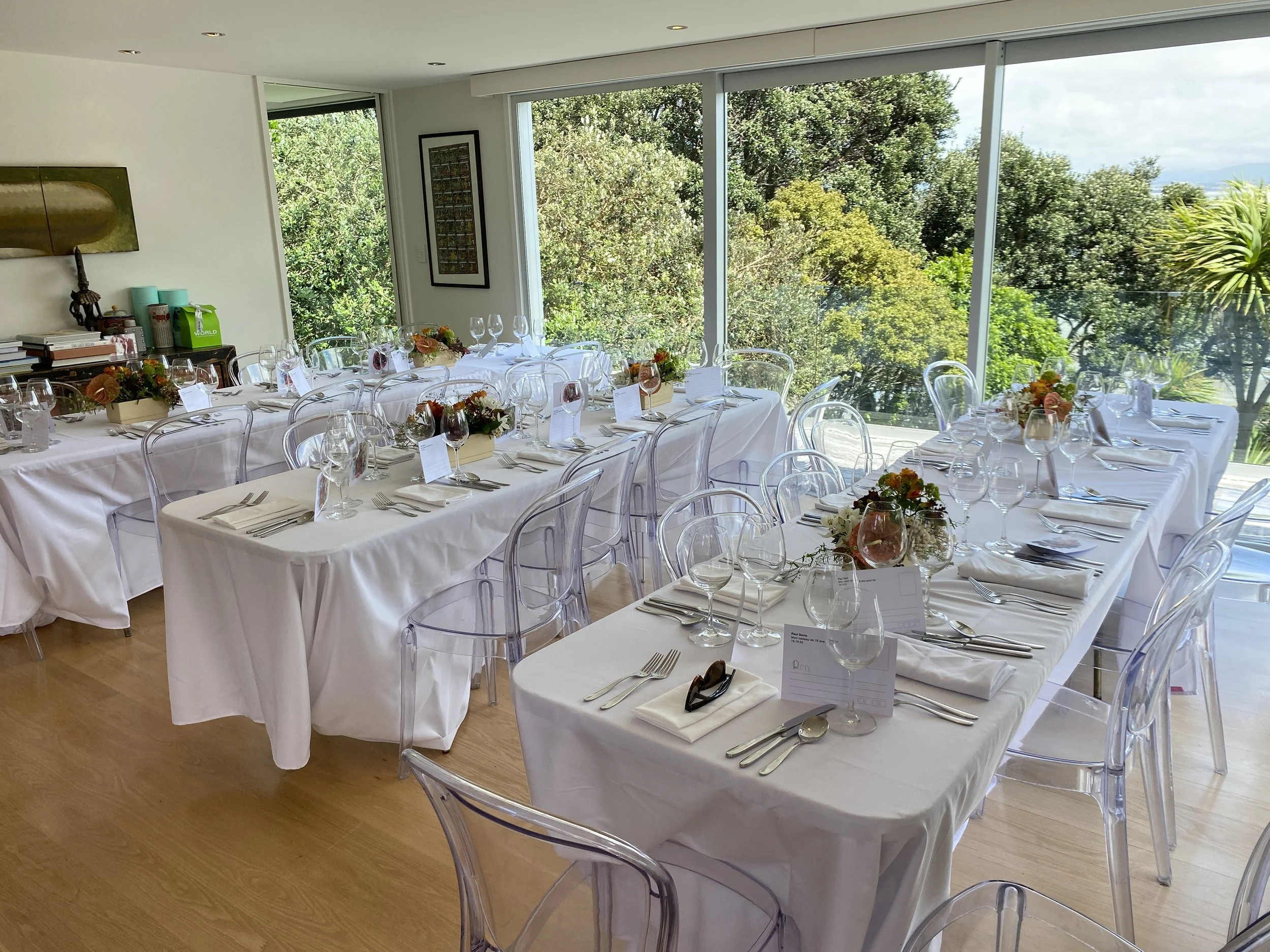 Dining area with elegantly set tables covered in white tablecloths, clear acrylic chairs, and floral centerpieces, next to large windows overlooking lush greenery.