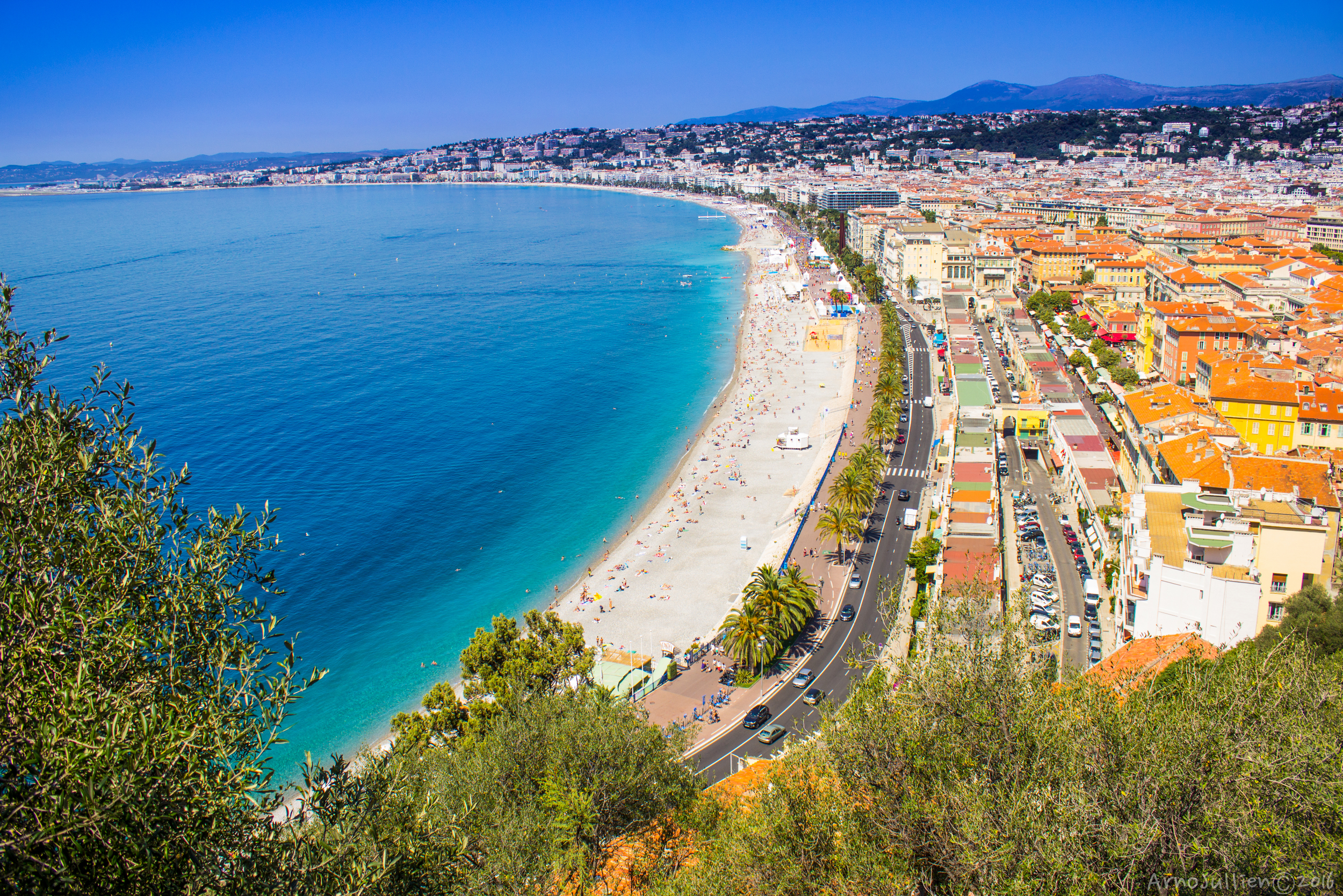 Panoramic view of Nice, France coastline with blue Mediterranean Sea, sandy beach, palm-lined Promenade des Anglais, and city buildings under a clear blue sky.