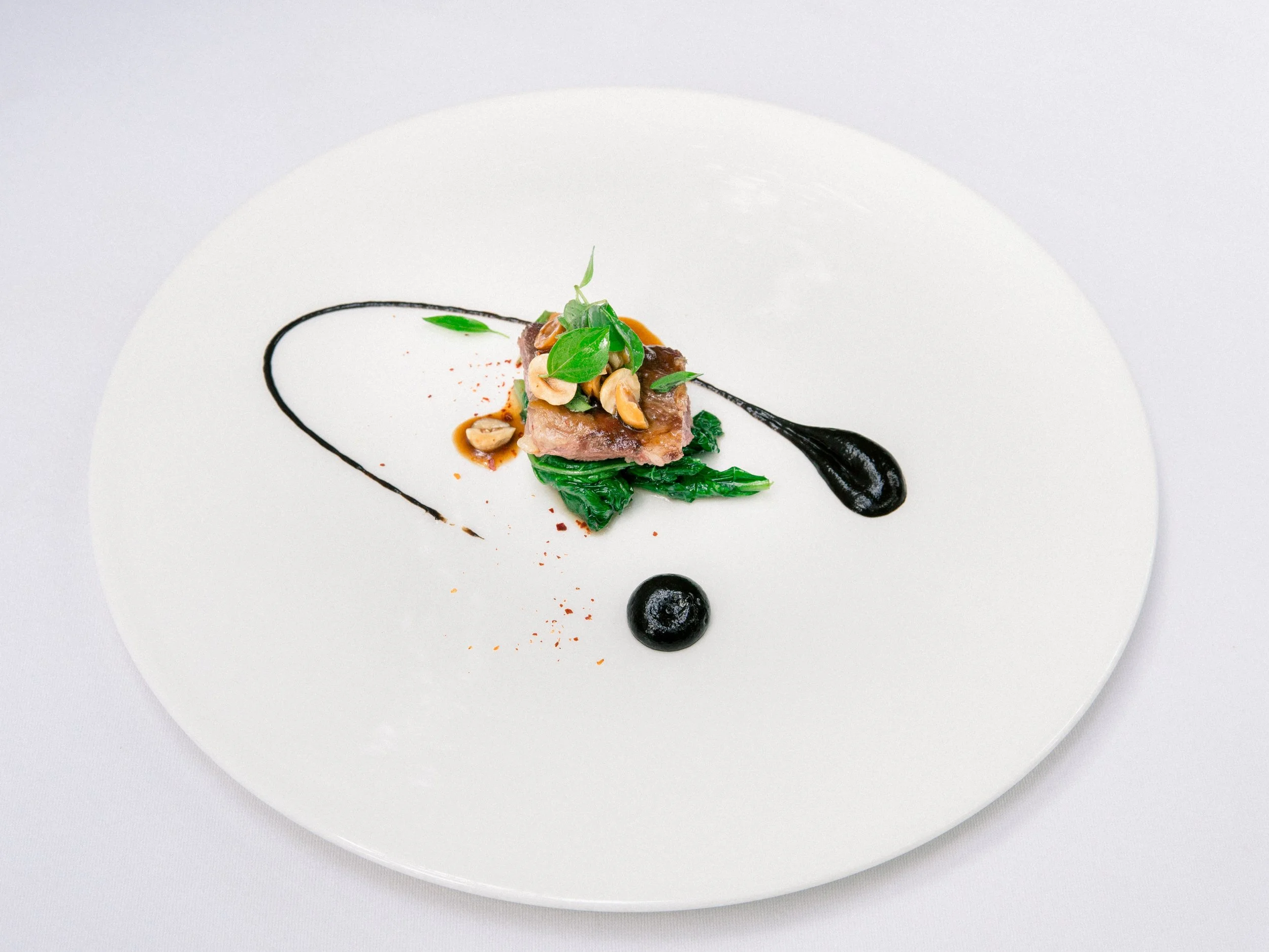 Gourmet dish with meat and greens on a white plate, garnished with black sauce and herbs.