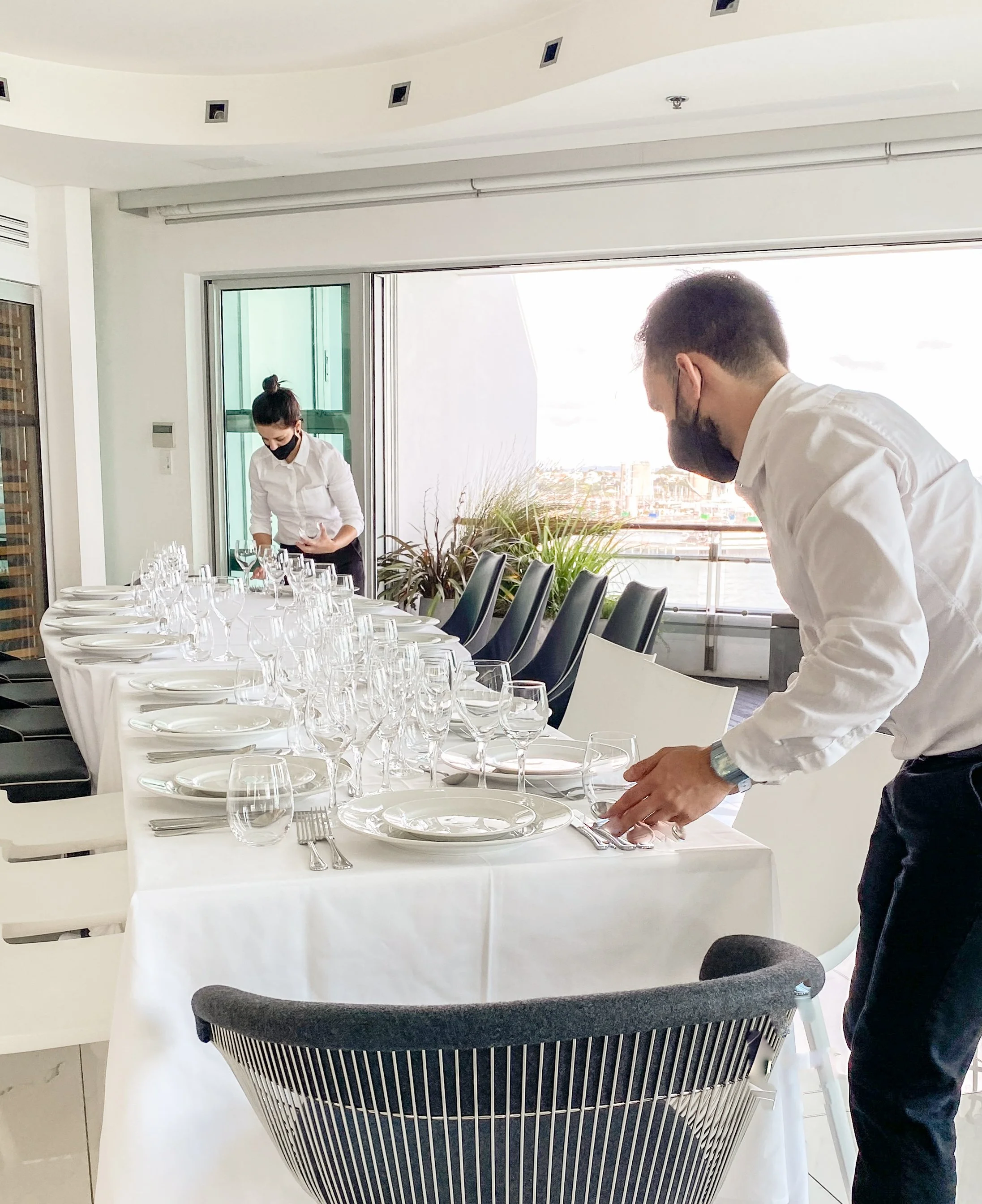 Two people setting a formal dining table with white tablecloth, plates, and glasses in a bright room with large windows.