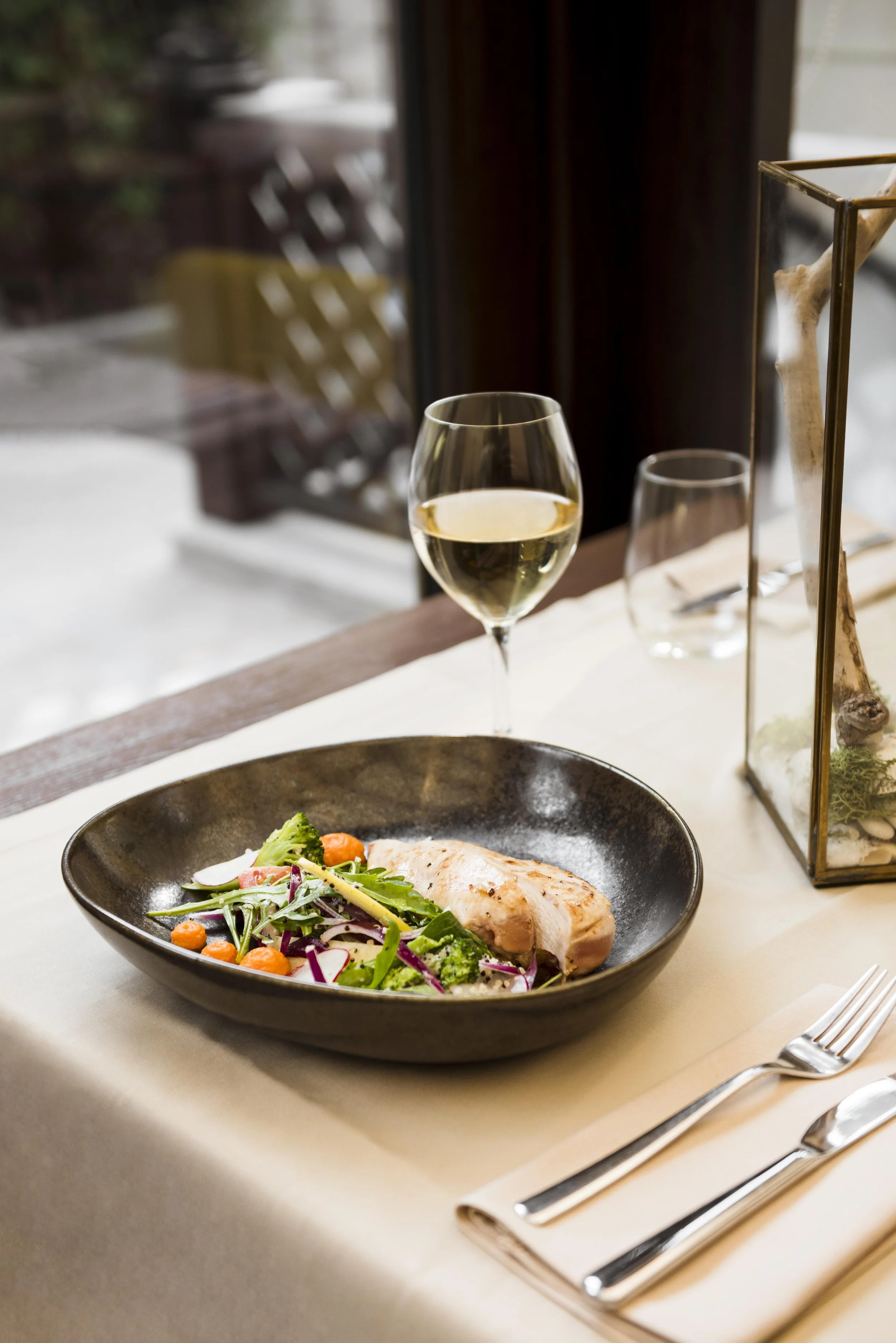 Plate with grilled chicken and salad on a table, glass of white wine, utensils, and a decorative lantern.