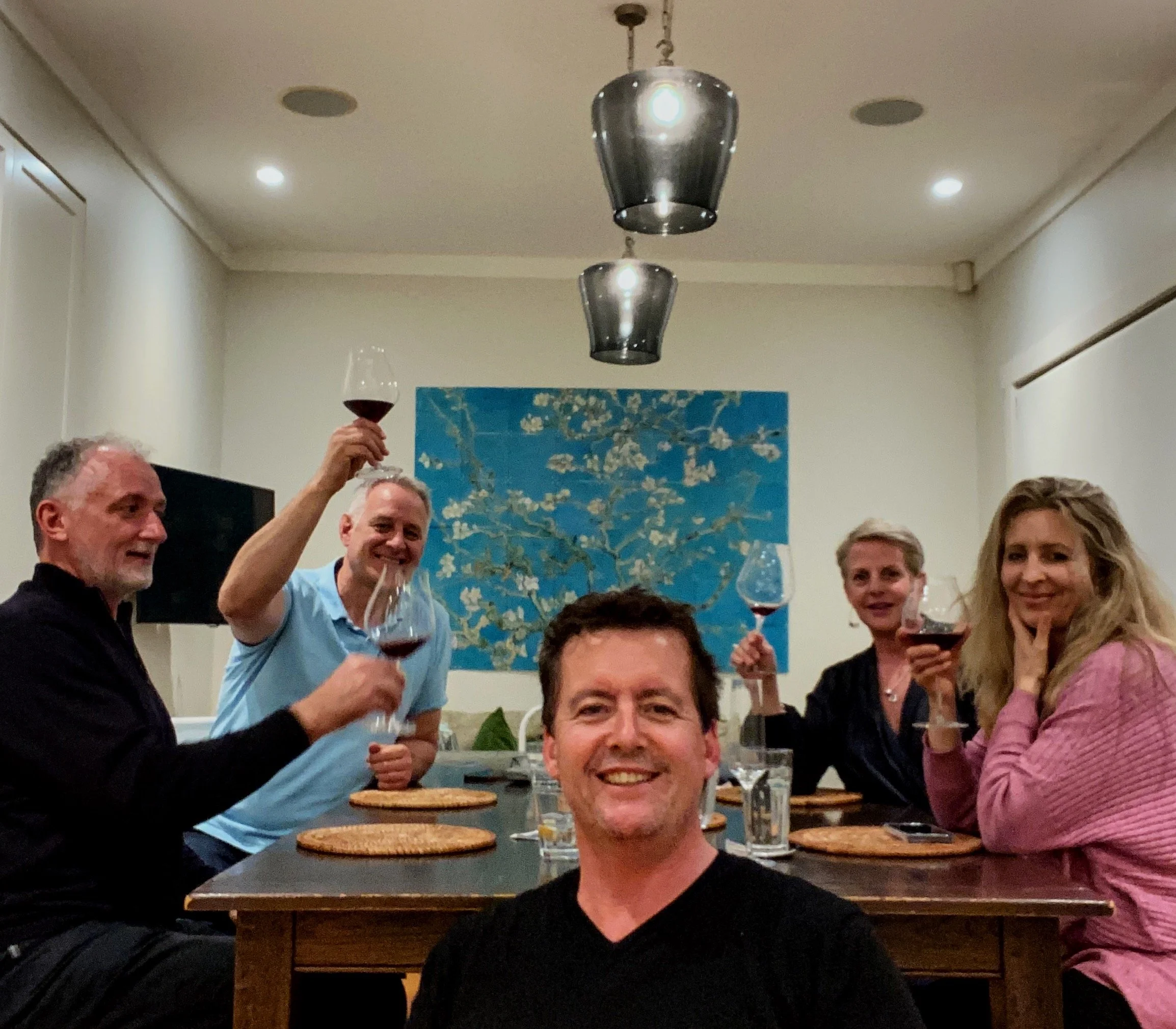 Group of five people sitting at a dining table, holding wine glasses, smiling, with a painting on the wall and hanging lights above.