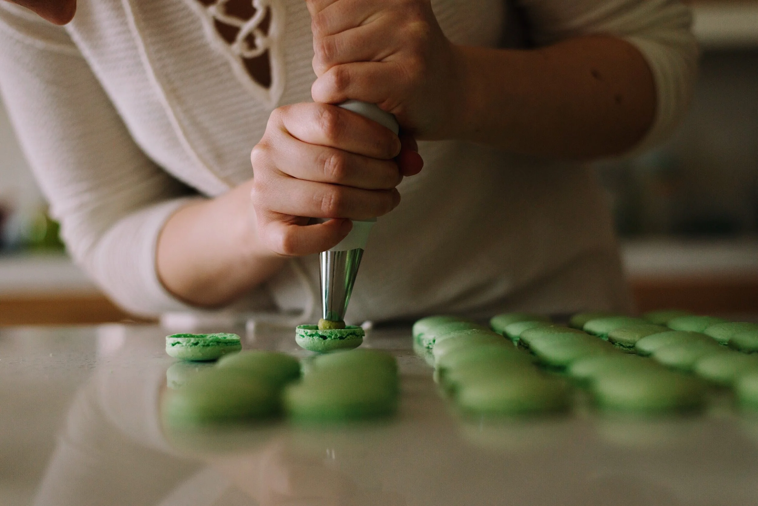 Person piping filling onto green macarons on a countertop.