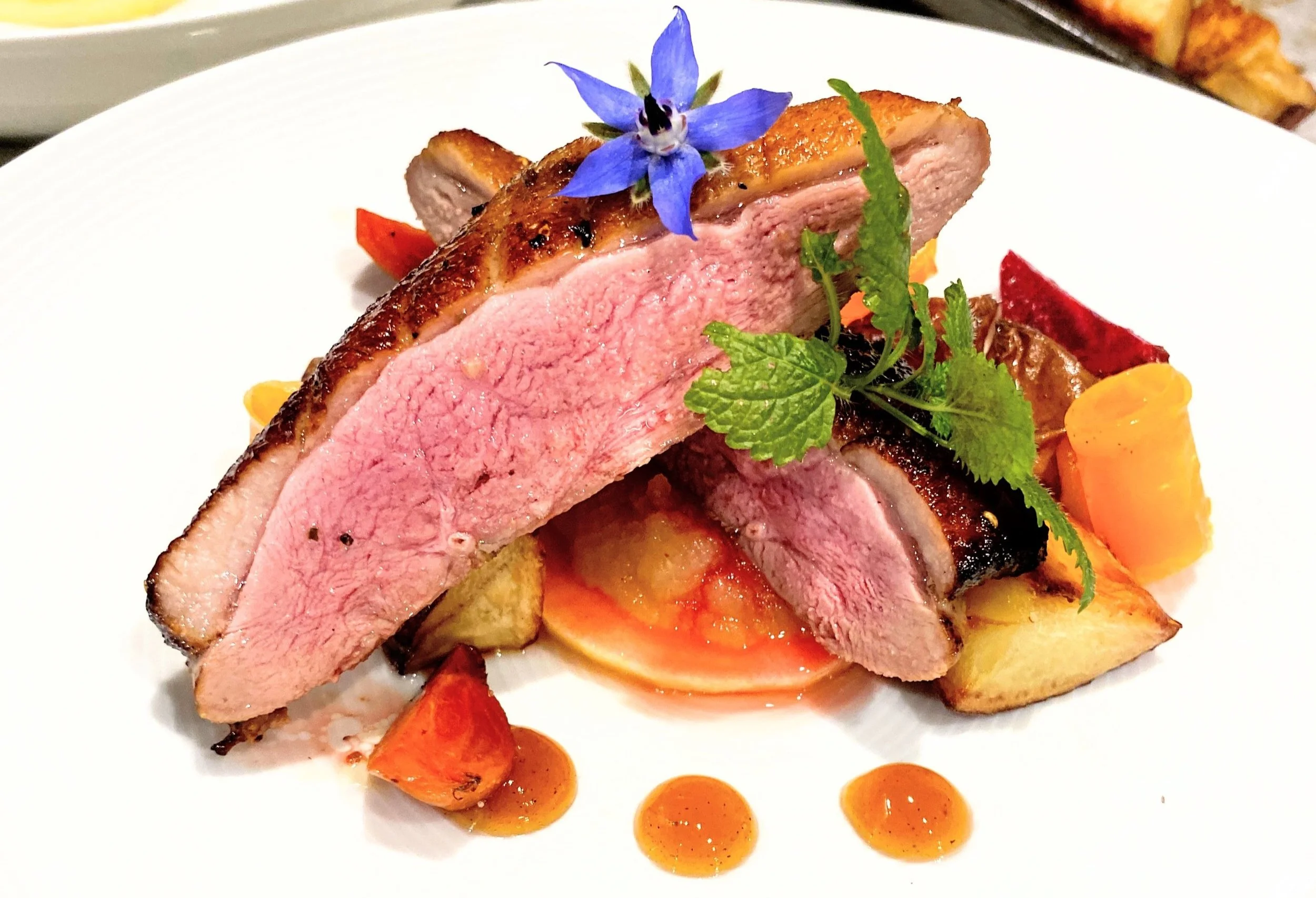 Sliced medium-rare duck breast garnished with a blue edible flower and fresh green herbs, served with roasted vegetables and sauce droplets on a white plate.