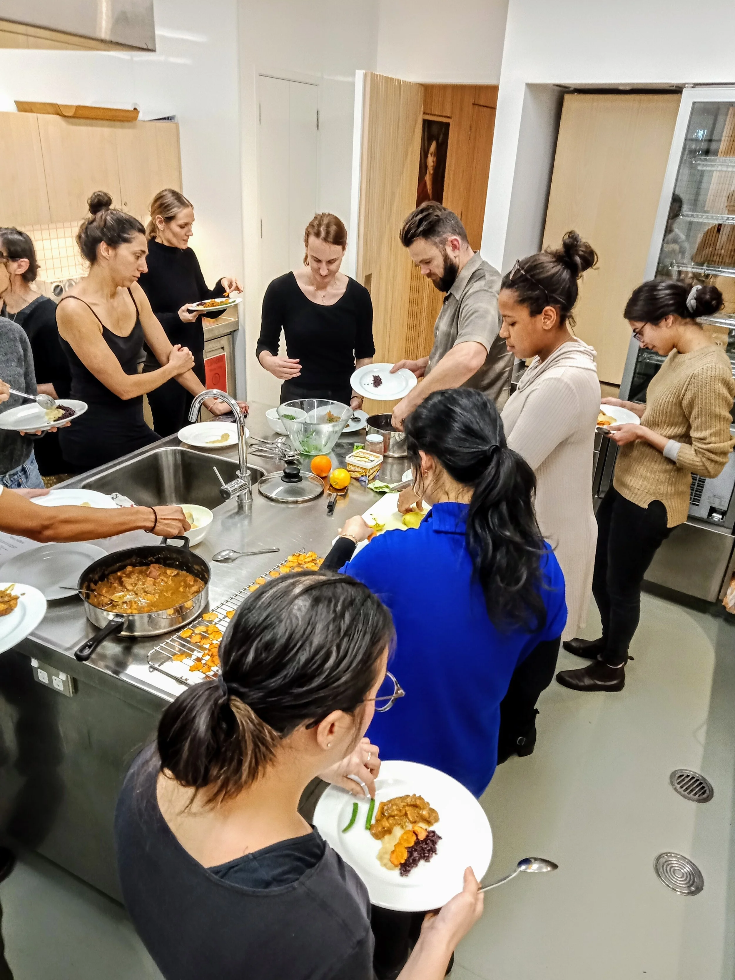 Group of people gathered in a kitchen serving food onto plates, with various dishes prepared on a countertop.