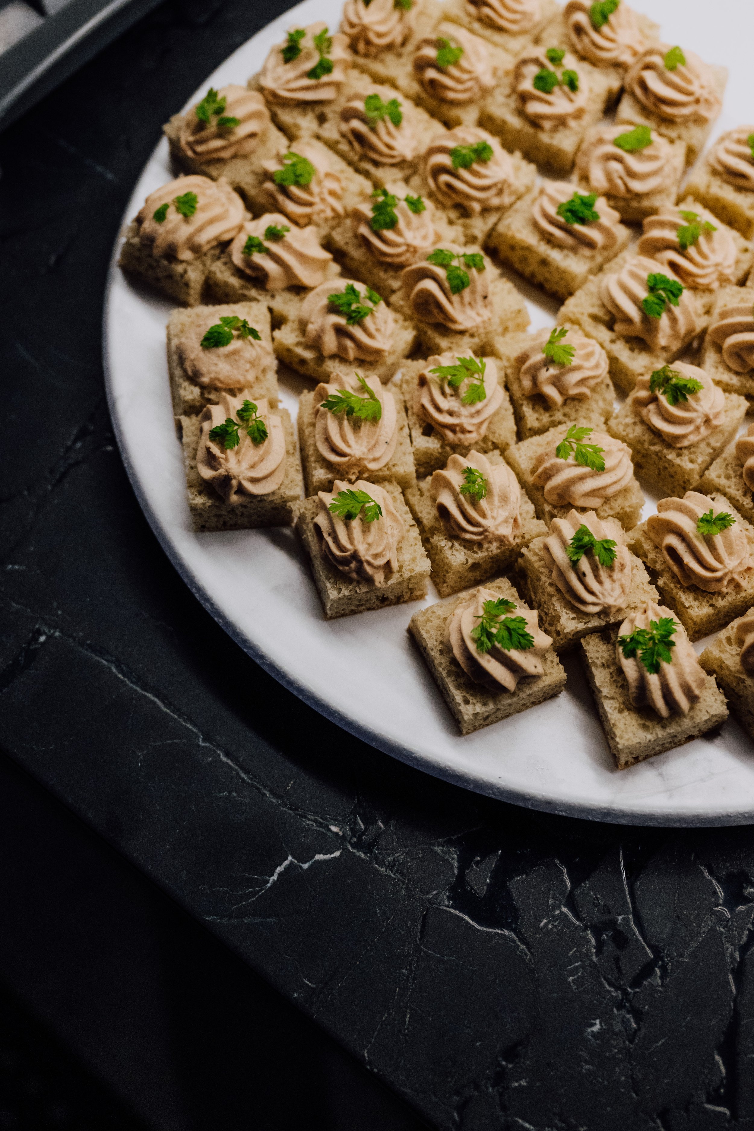 Close-up of a plate with small square appetizers, featuring bread topped with a creamy spread and garnished with parsley, placed on a dark textured surface.