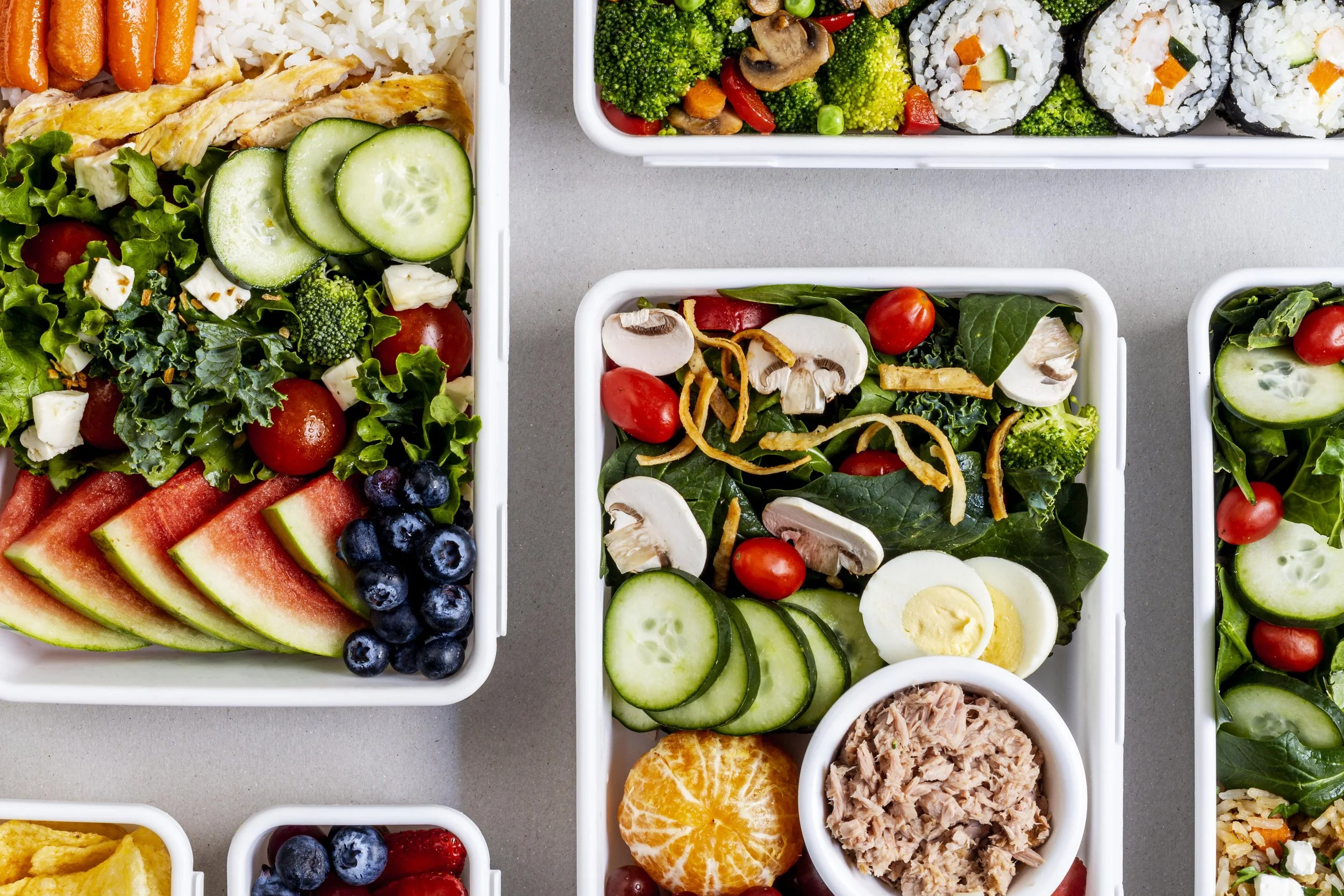 Multiple containers of fresh salads with ingredients like cucumbers, cherry tomatoes, broccoli, mushrooms, boiled eggs, blueberries, watermelon, mandarin orange, and tuna on a gray surface.