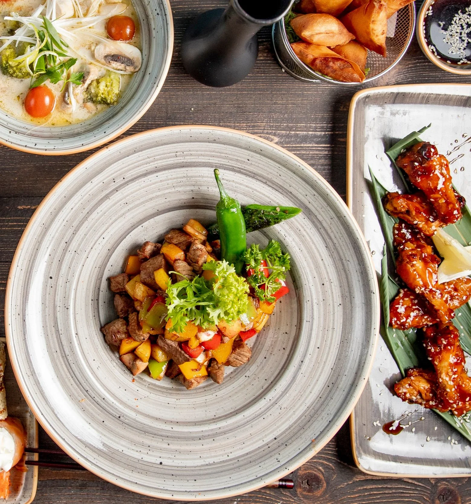 Assorted Asian dishes including stir-fried beef with vegetables, two types of glazed chicken wings, a bowl of soup with vegetables and cherry tomatoes, and fried spring rolls on a dark wooden table.