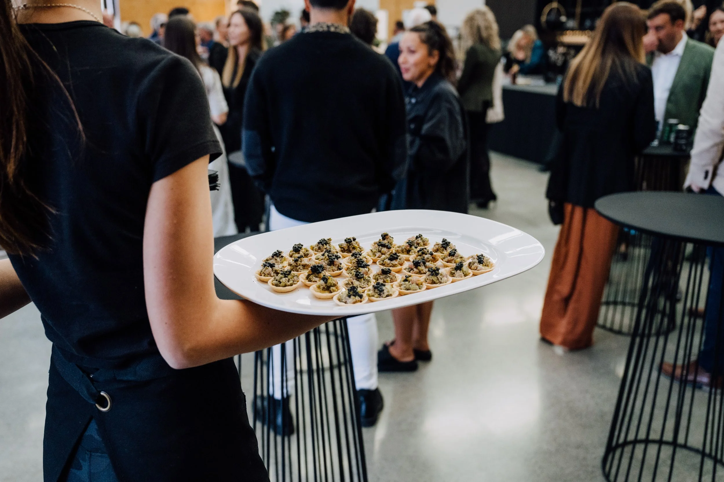 A person in black holding a white tray with small appetizers topped with black caviar at a crowded social event.