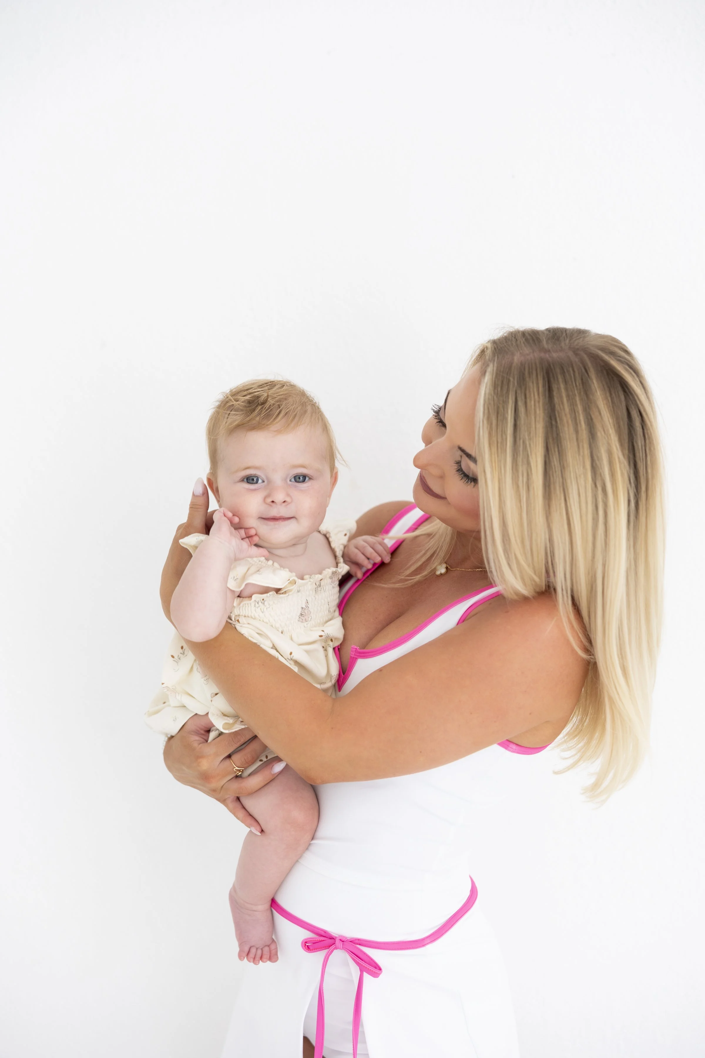 A woman with long blonde hair holding a young toddler girl with red hair, both smiling, against a plain white background.