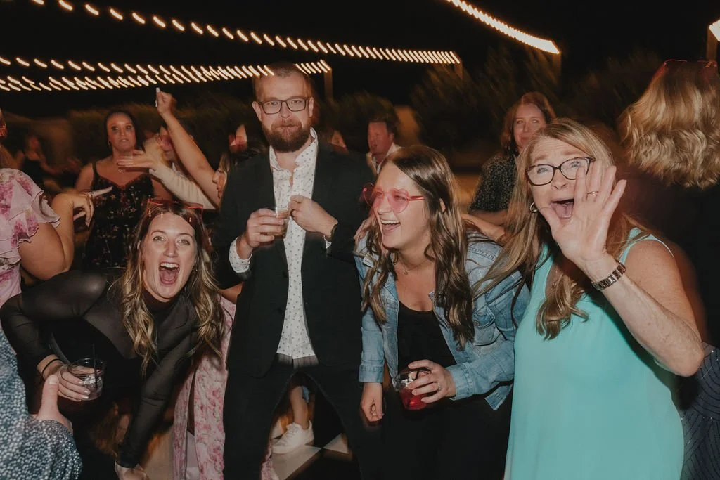 People celebrating at an outdoor party, some wearing glasses and holding drinks, with string lights overhead.