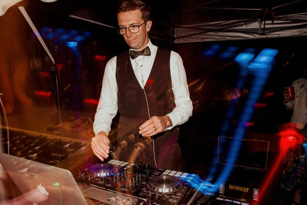 DJ wearing a suit with a bow tie, using DJ equipment in a dimly lit setting with colorful lights.