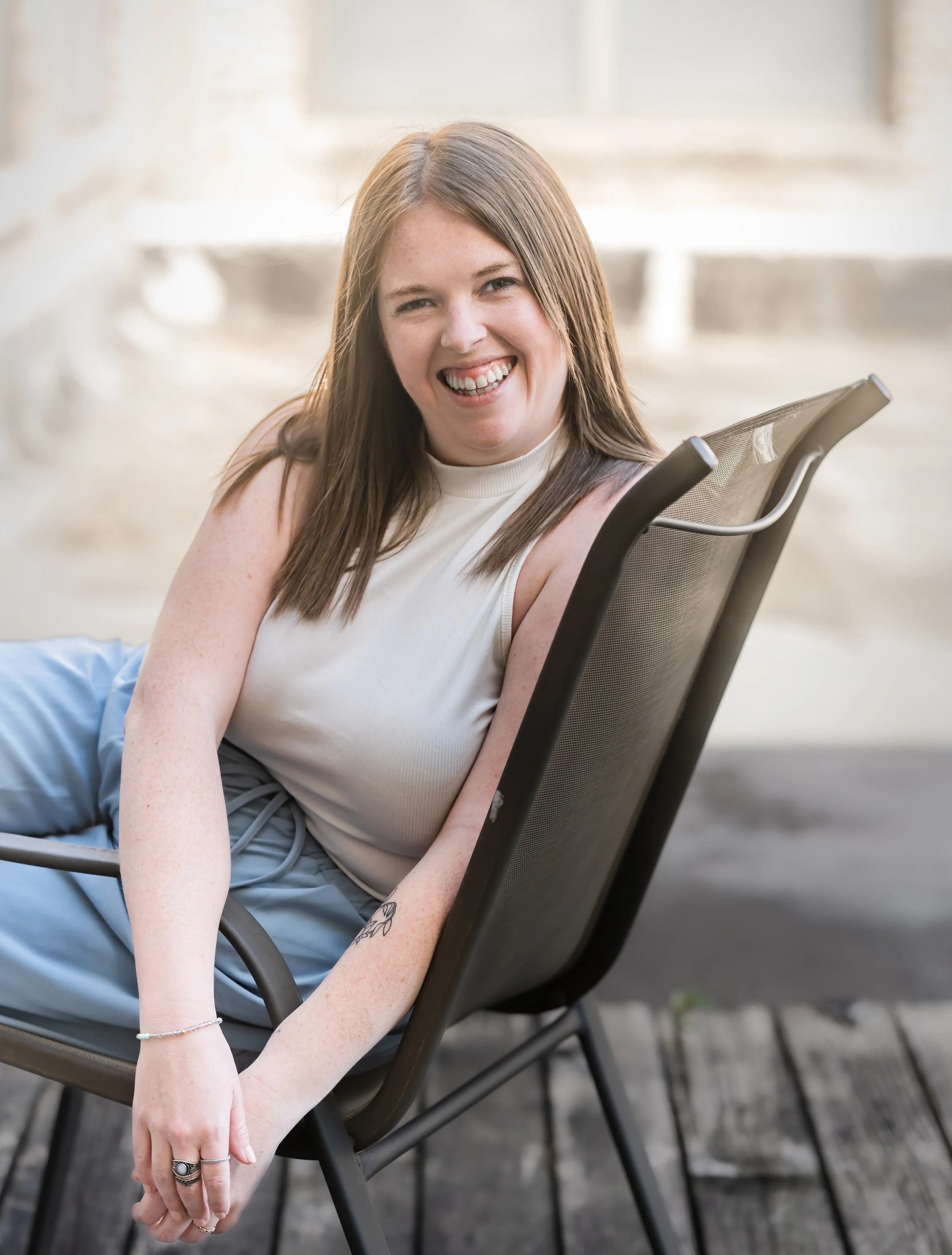 A young woman with light skin and brown hair sitting on a black outdoor chair, smiling and looking at the camera, with a blurred stone or concrete background.