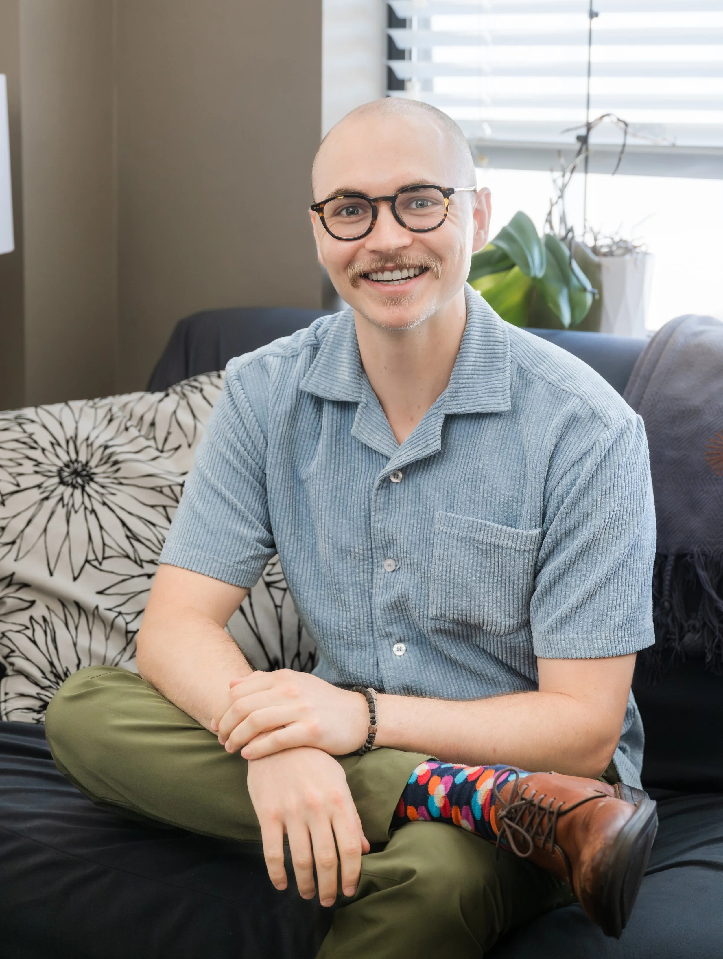 A young man with glasses, a mustache, and a bald head sitting on a black couch, smiling, wearing a light blue shirt, olive green pants, colorful socks, and brown shoes in a well-lit room with curtains and houseplants in the background.