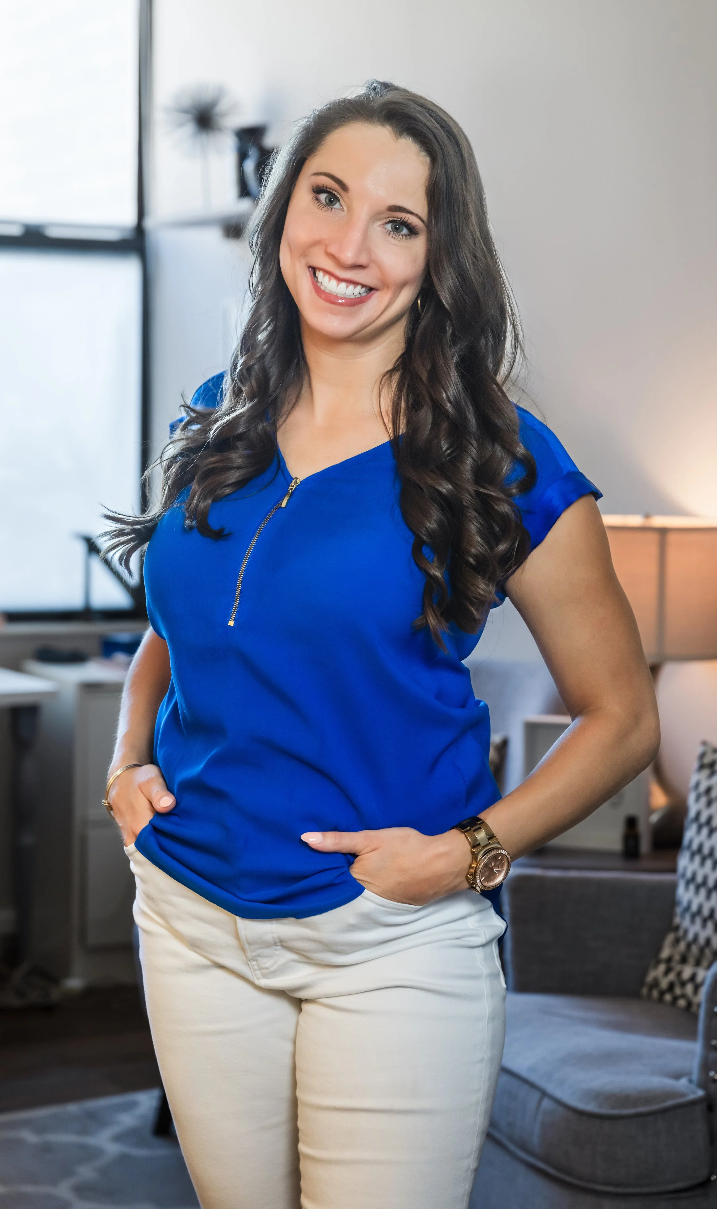 A woman with long, wavy dark hair, wearing a bright blue top with a zipper, beige pants, and a gold watch, standing in a modern, well-lit living room with a sofa and lamp in the background.