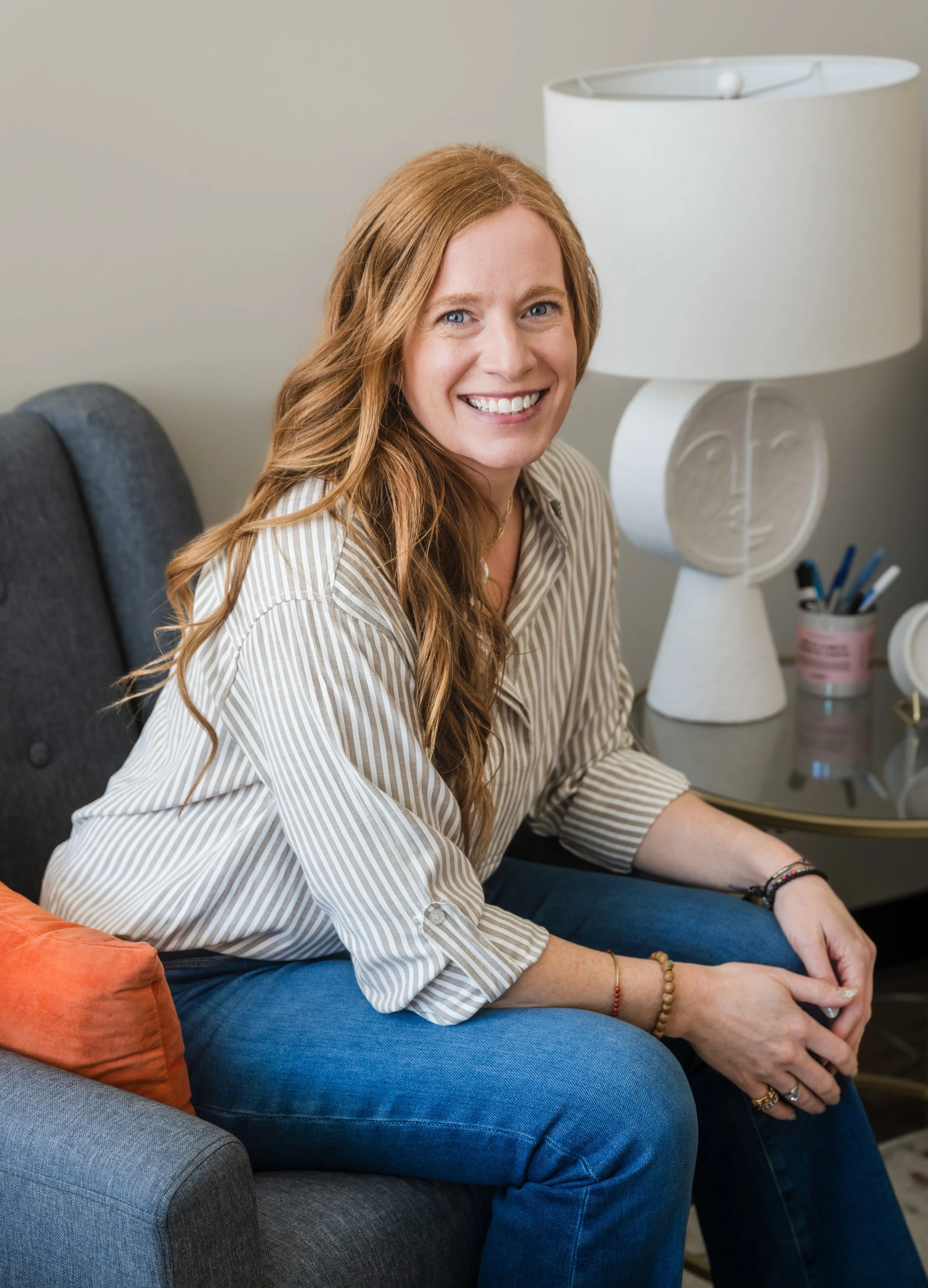 A woman with long red hair smiling and sitting on a gray couch, next to a side table with a lamp and decorative items.