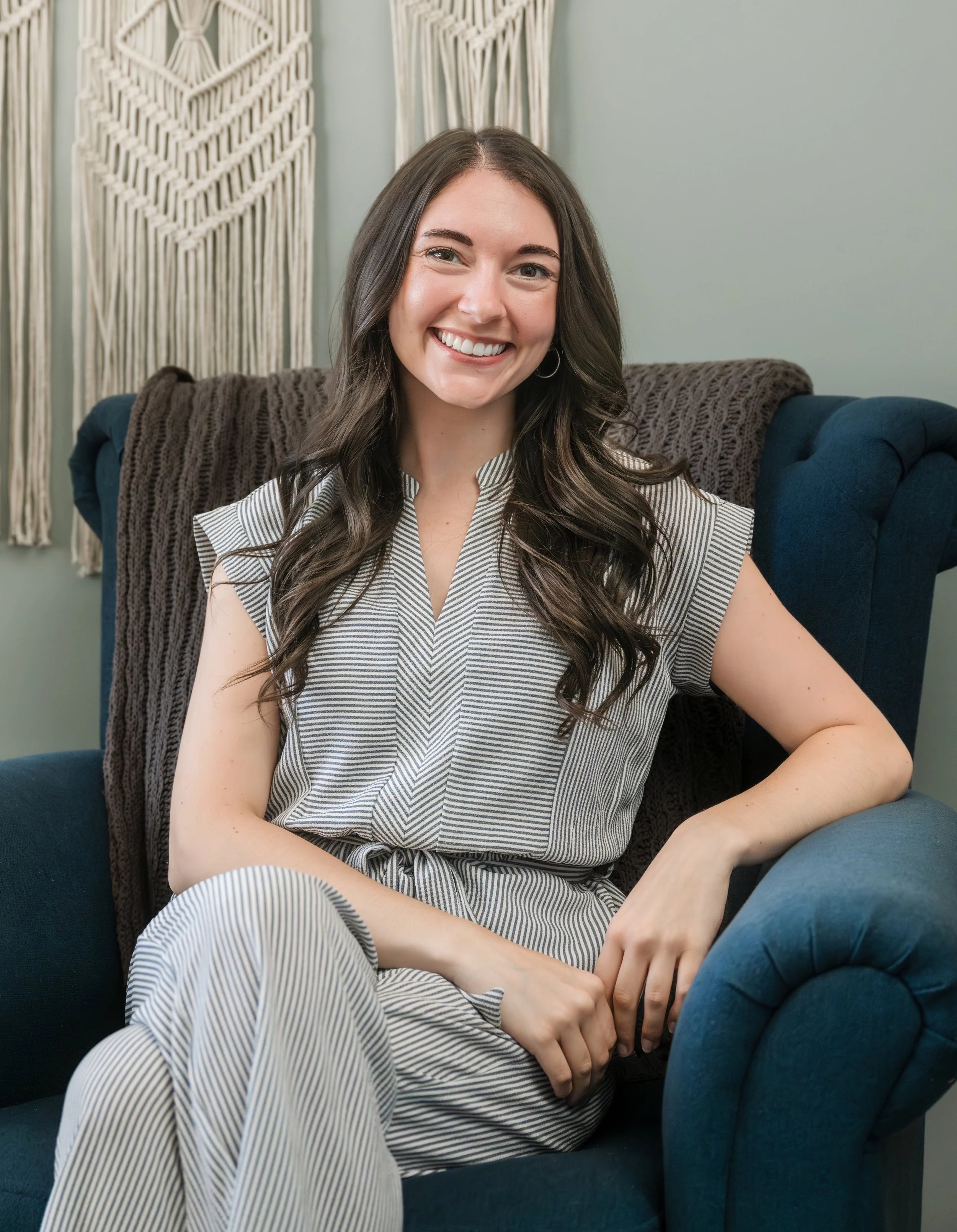 A woman with long dark hair, smiling, sitting in a blue armchair with a dark brown blanket, in a room decorated with textile wall hangings.