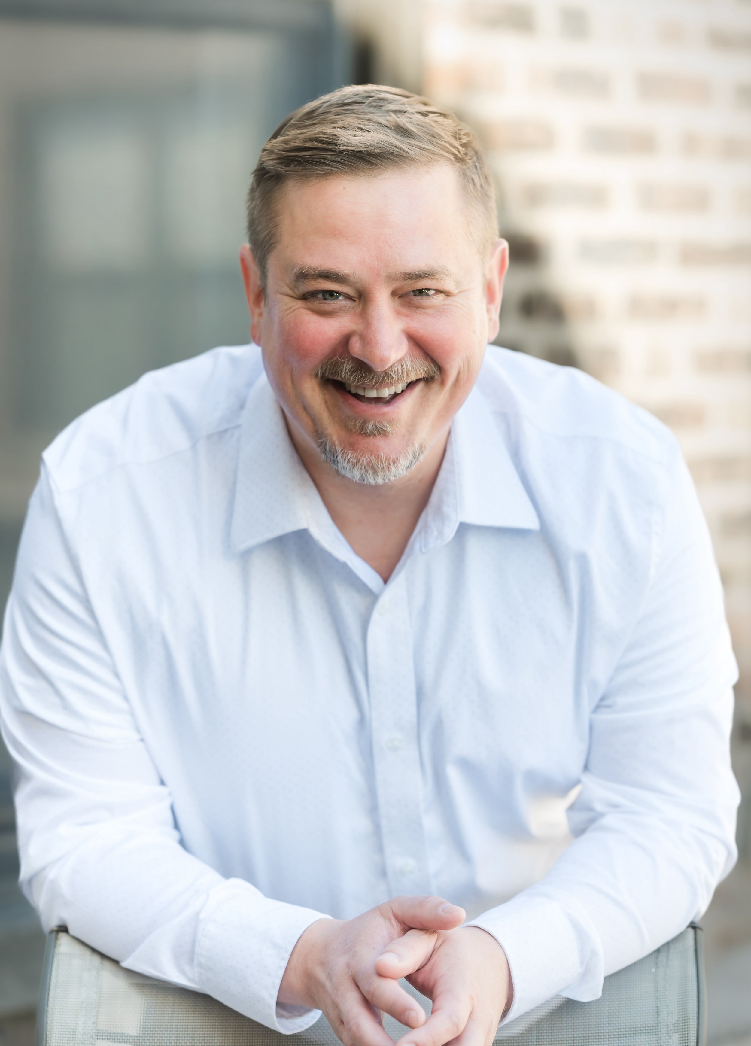 Smiling man with light brown hair, beard, and mustache, wearing a white button-up shirt, sitting outdoors with a blurred cityscape background.