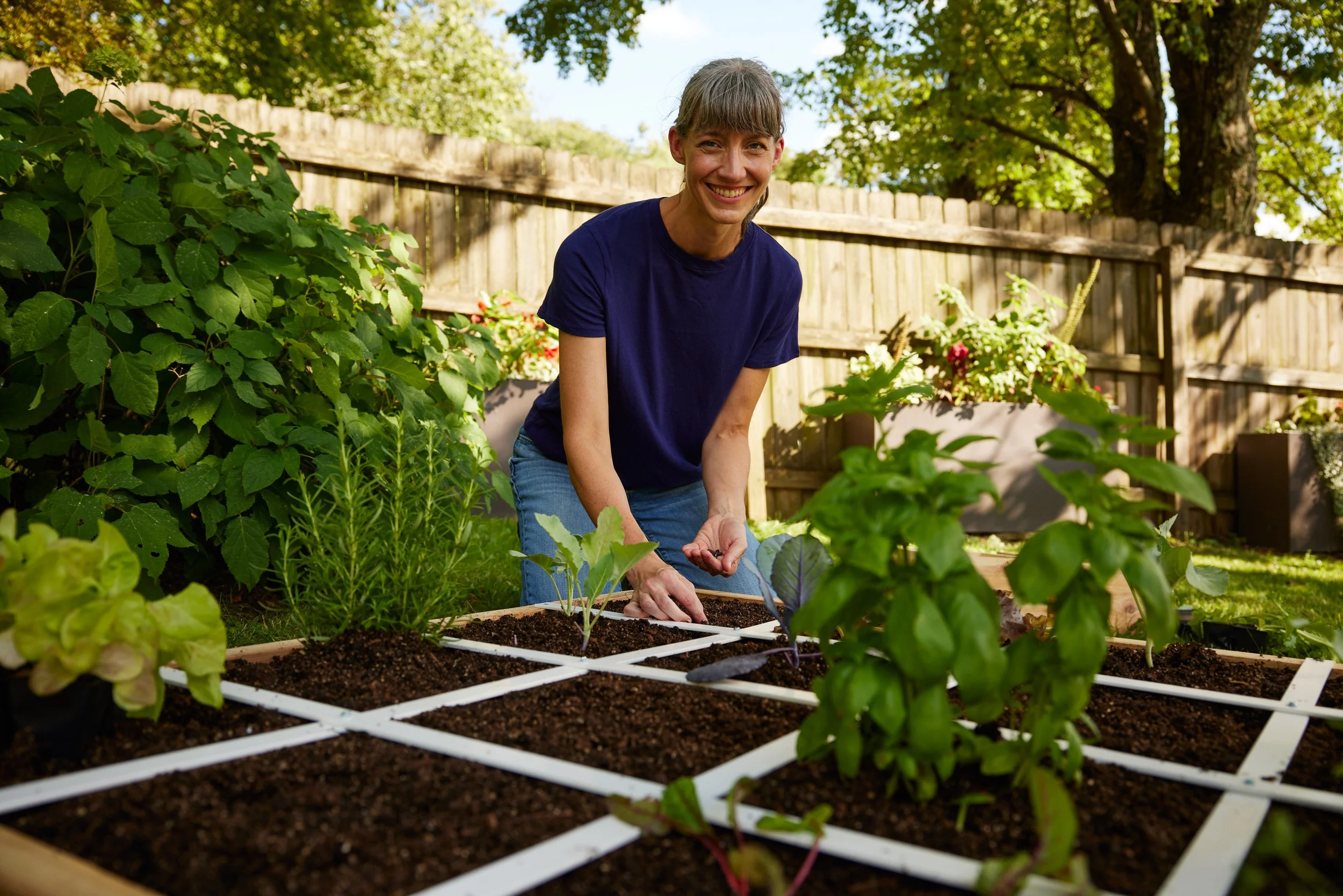 A woman with gray hair tied back, wearing a navy blue t-shirt and jeans, gardening in a backyard during daytime. She is smiling and planting small seedlings in a raised garden bed with dark soil, surrounded by green plants and a wooden fence, with tr