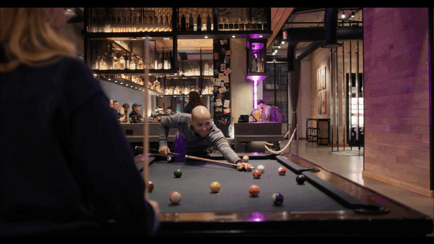 Person playing pool in a dimly lit bar with bottles on shelves and patrons in the background.